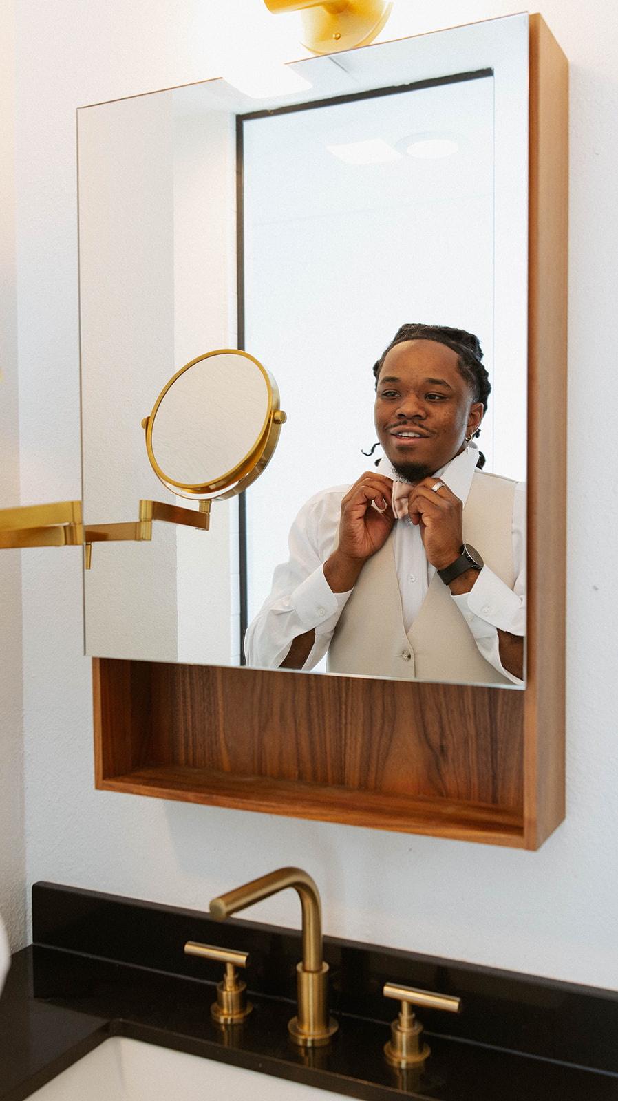 Groom adjusting his bow tie in the mirror before the ceremony, wearing a beige vest and white shirt in a softly lit bathroom.