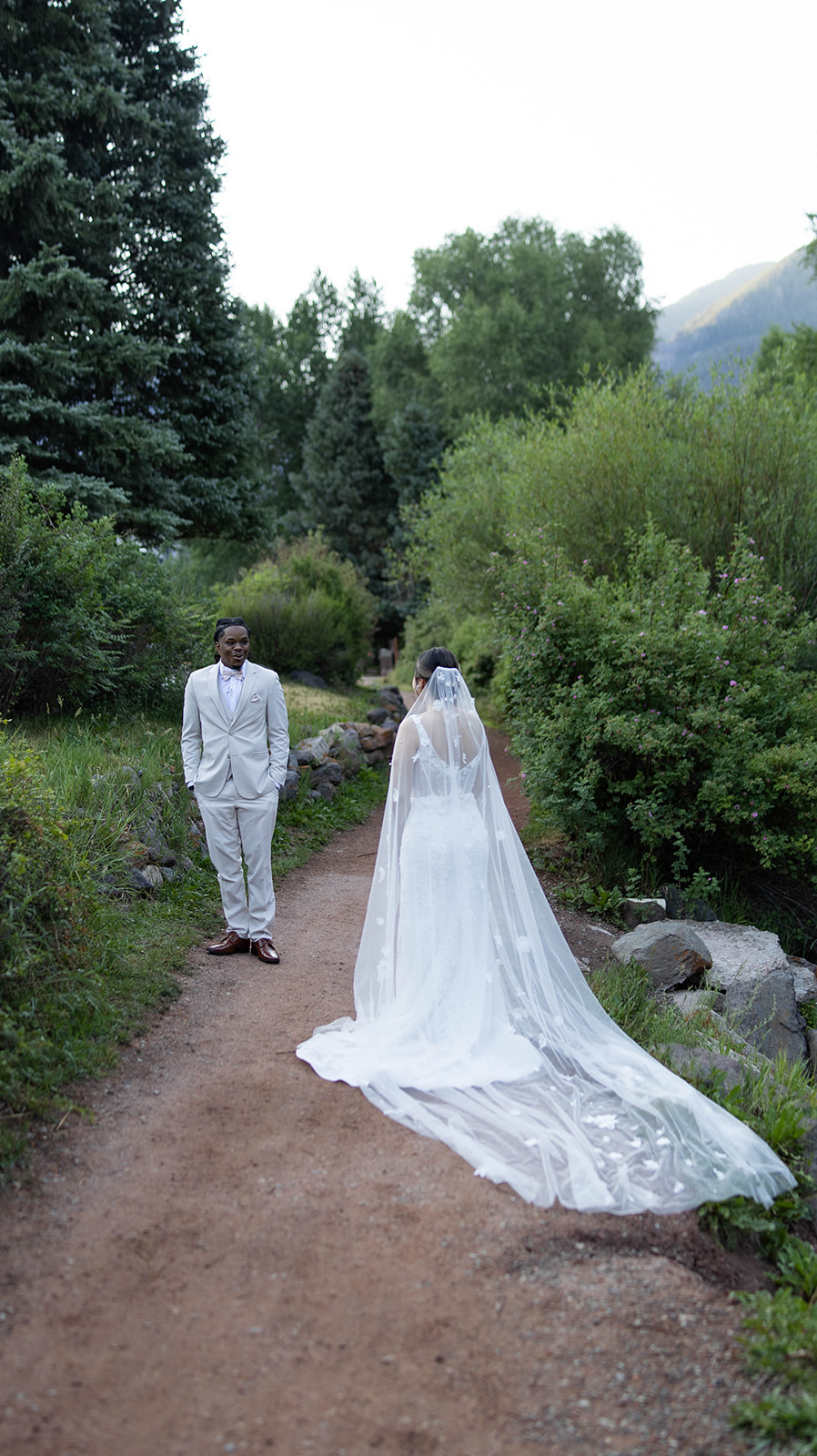 Bride in a long, floral-detailed veil walking toward her groom on a forested path surrounded by tall pines during their Telluride elopement.