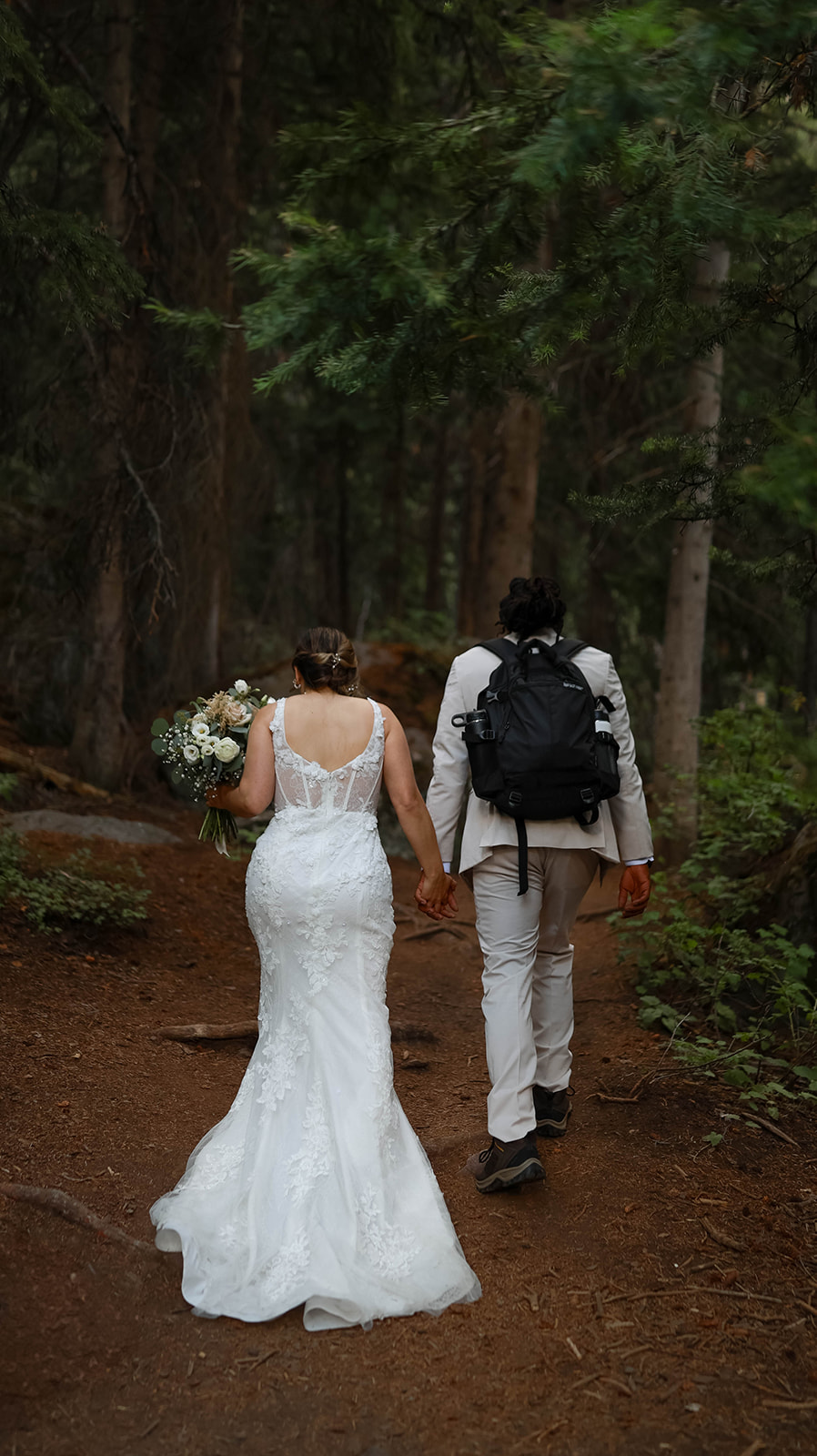 Couple walking hand in hand along a wooded trail, the groom wearing a hiking backpack and the bride carrying a bouquet.