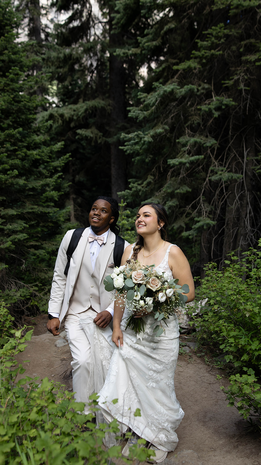 Bride and groom walking through a lush forest trail, holding hands and smiling, bride carrying a bouquet of white and blush flowers for her Telluride elopement.