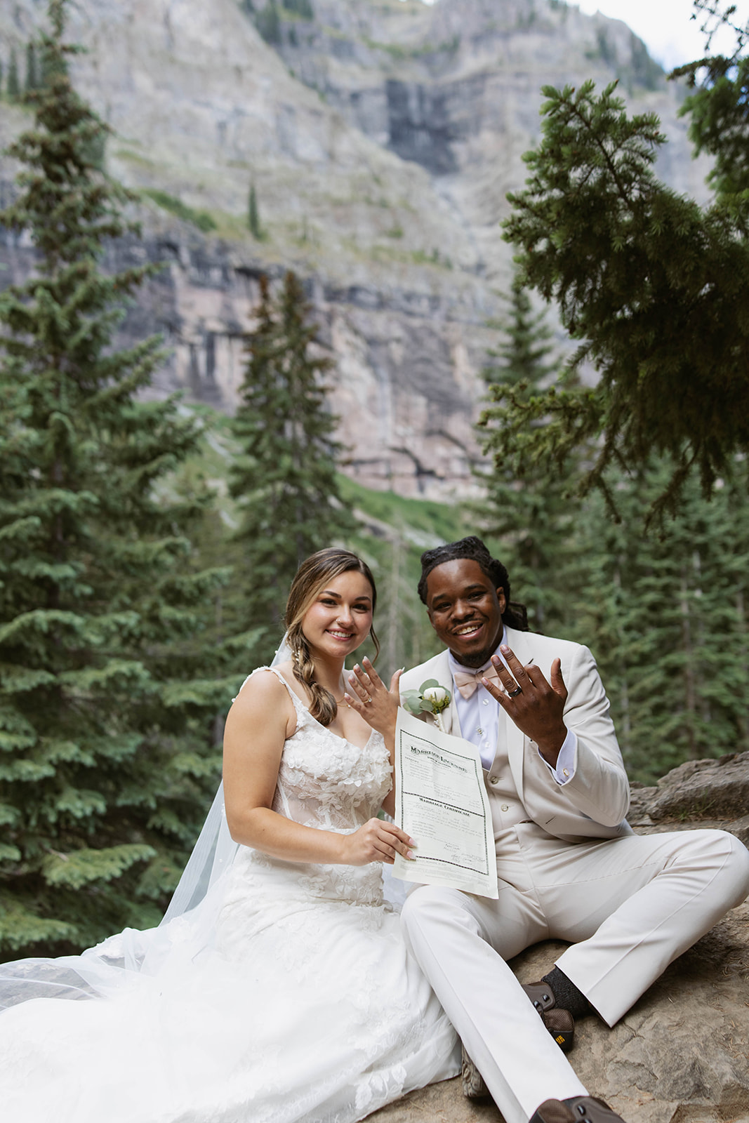 Newlyweds sitting on a rock surrounded by pine trees, proudly showing their wedding rings and holding their marriage license.