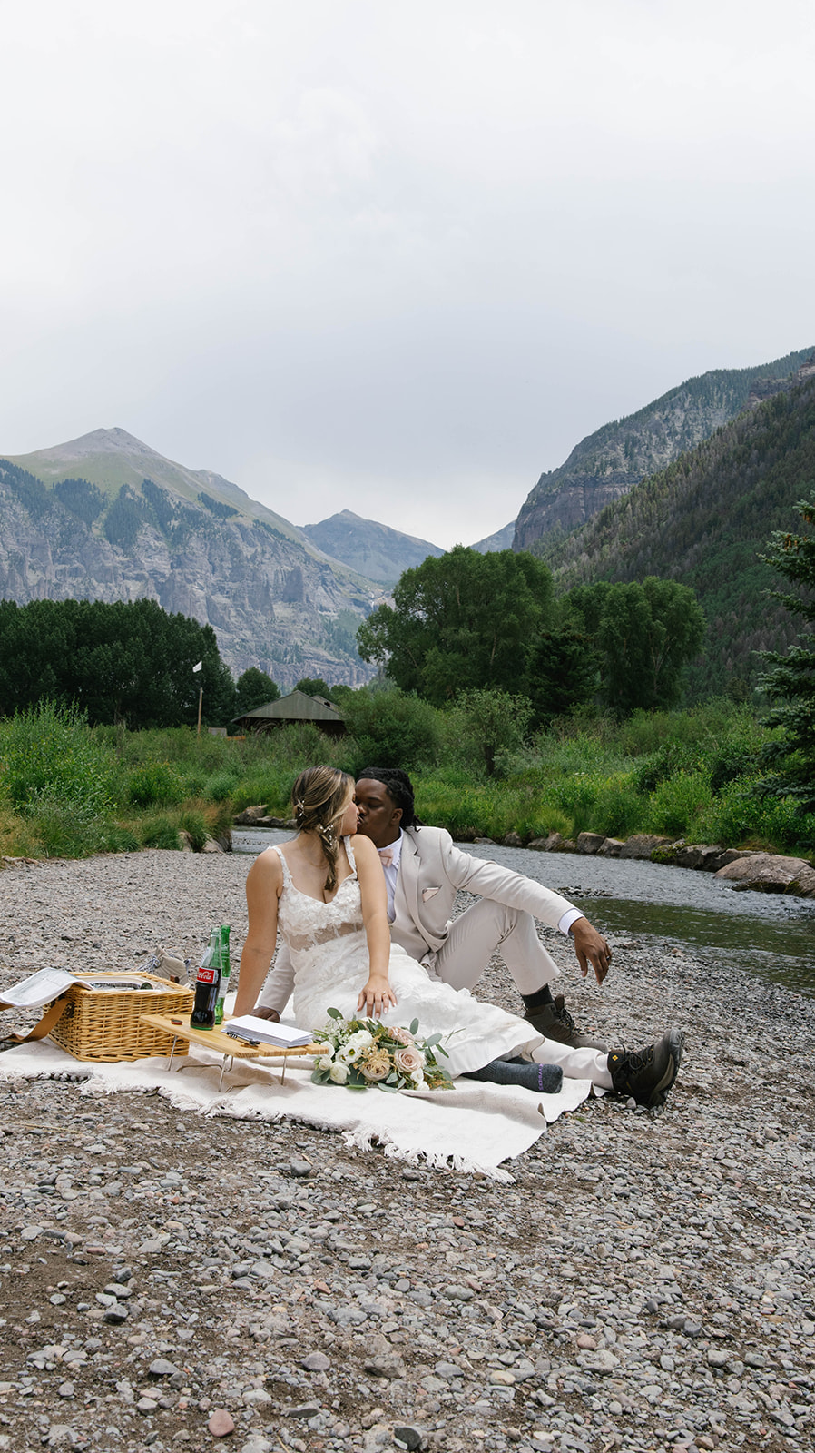 Couple enjoying a picnic by a mountain stream, sitting on a blanket with flowers, snacks, and vintage soda bottles. 