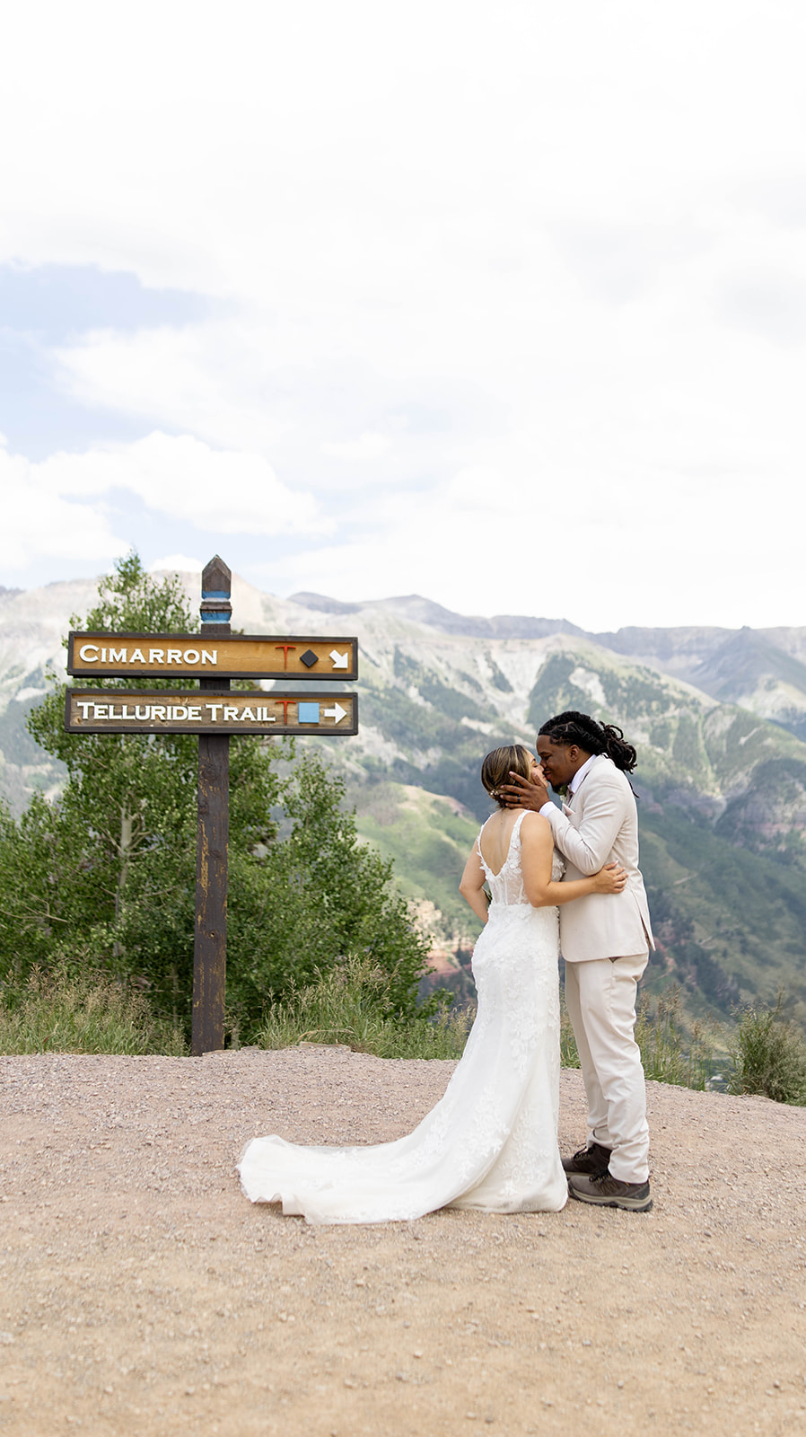 Bride and groom share a kiss at the top of a mountain trail, standing beside a wooden sign pointing to Cimarron and Telluride Trail, with sweeping alpine peaks in the background after their Telluride elopement.