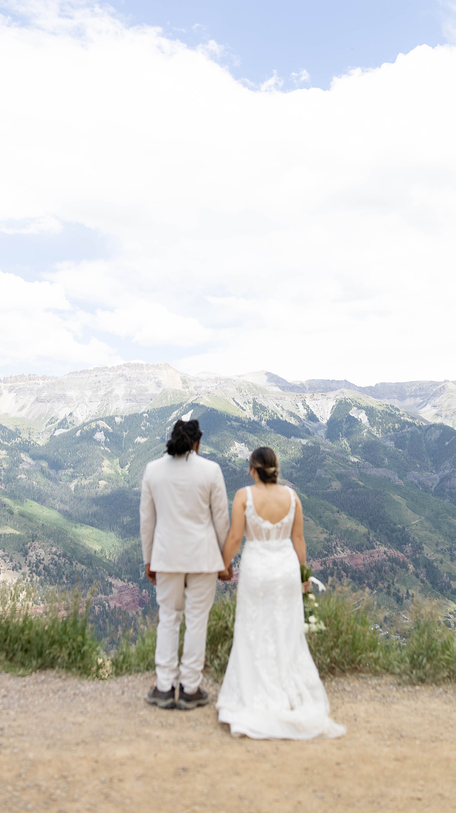 Couple standing hand in hand, facing sweeping mountain views under a partly cloudy sky during their Telluride elopement, the bride in a lace gown and the groom in a light suit.