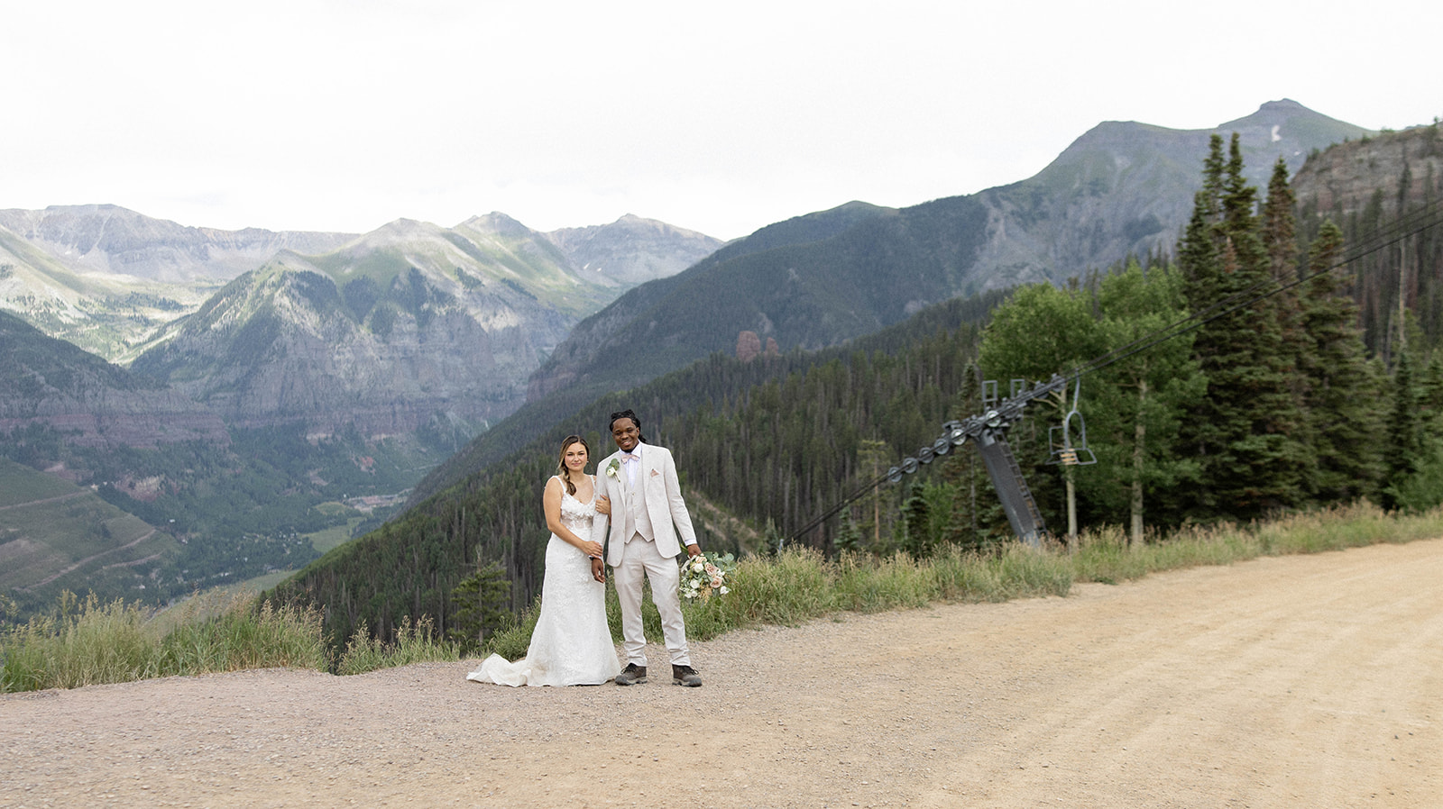 Bride and groom stand hand in hand on a dirt road surrounded by pine trees and the San Juan Mountains.