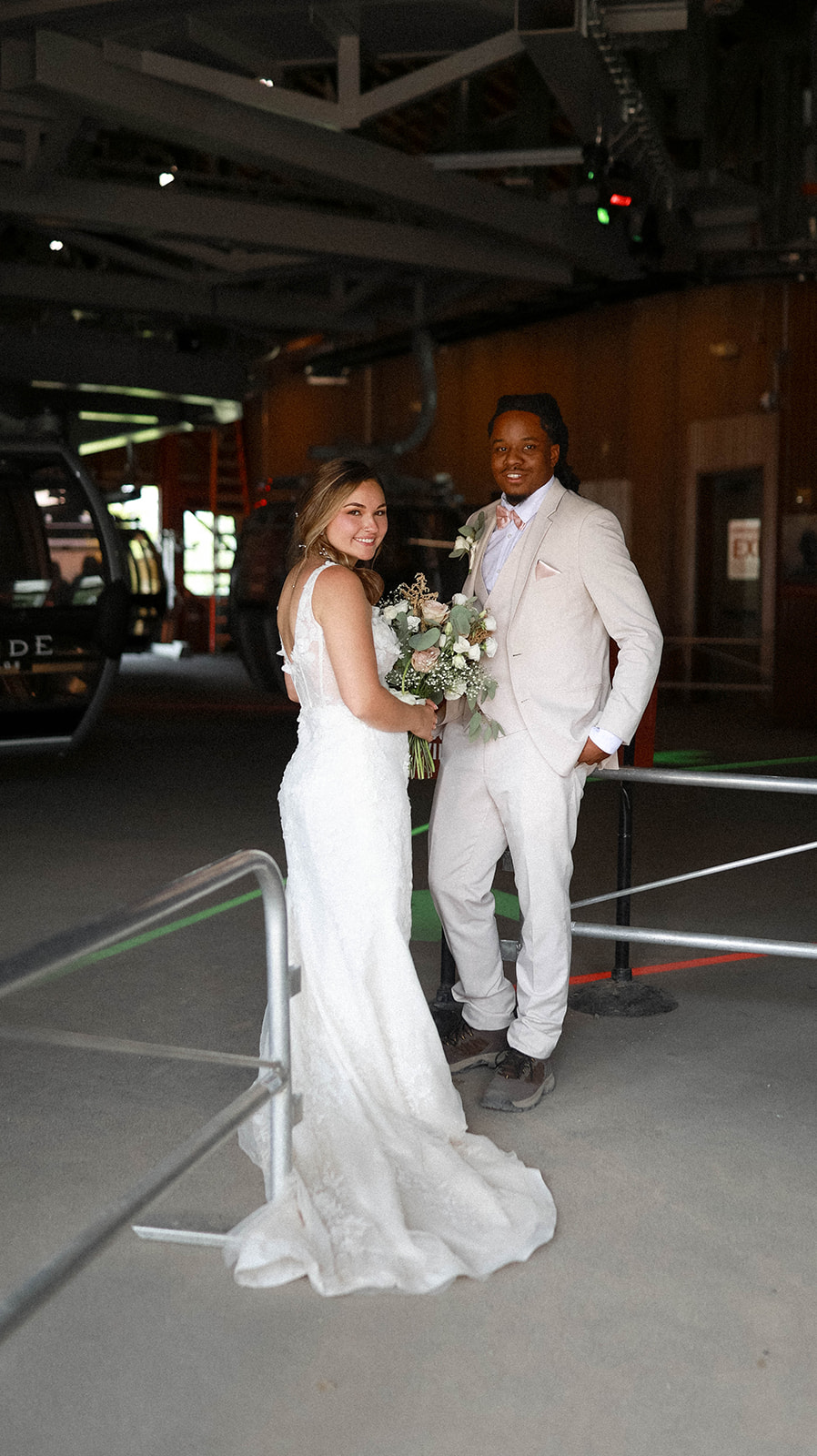 Bride and groom smiling while standing inside a gondola station, holding a bouquet of white and blush flowers.