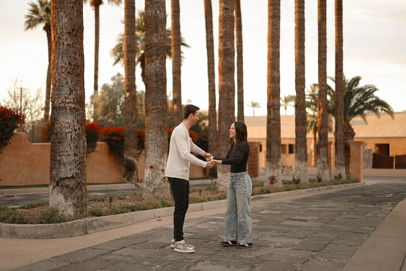 A couple stands hand in hand, smiling at each other in a palm tree–lined courtyard.