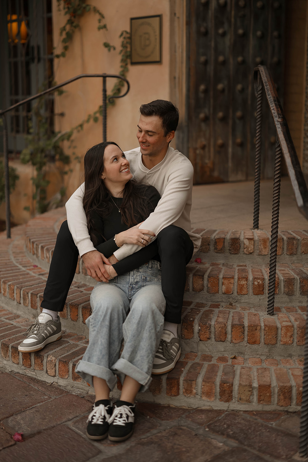A couple sits on brick steps, holding each other and smiling while leaning close.