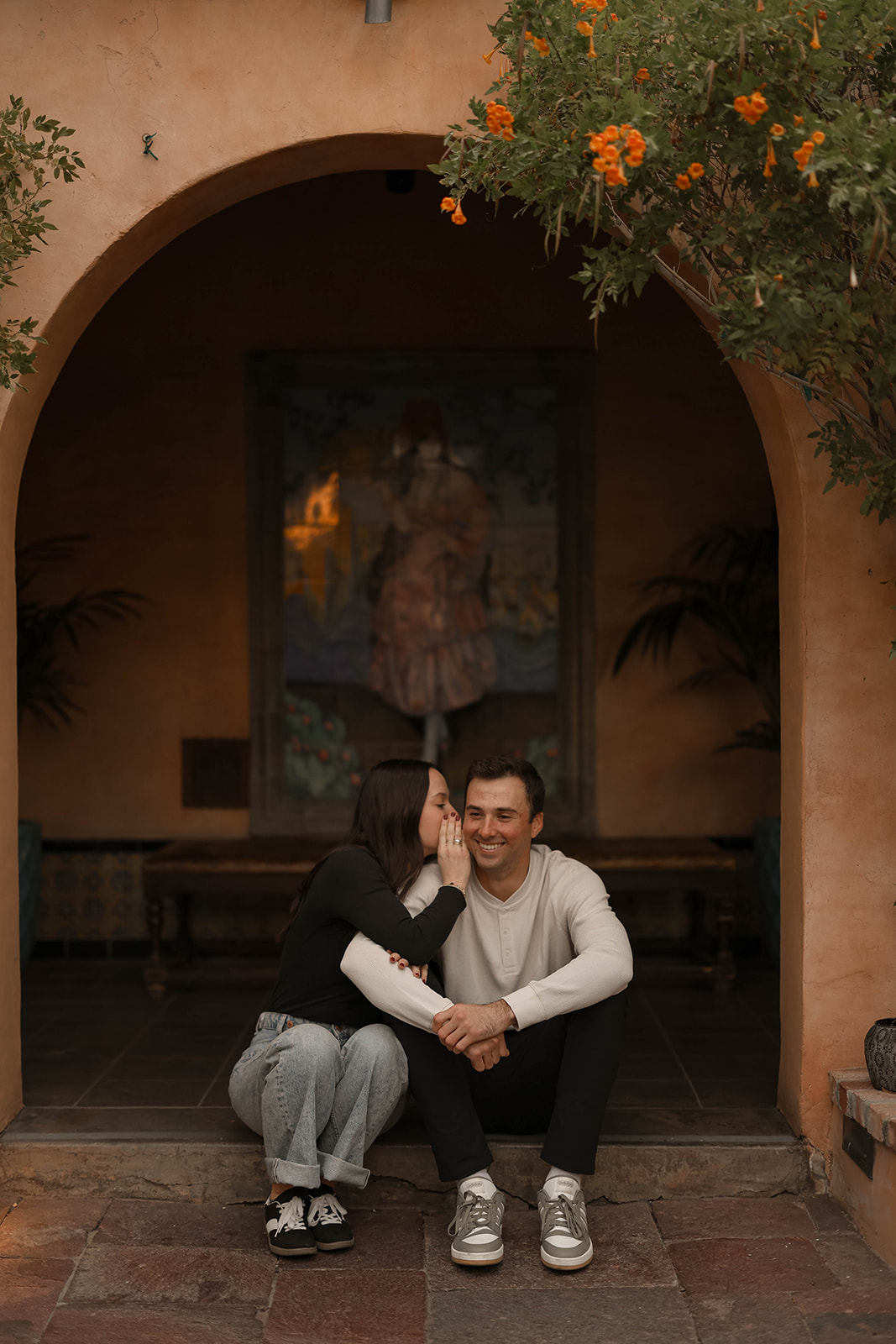 A woman whispers into her partner’s ear as they sit together under a warm-toned archway.