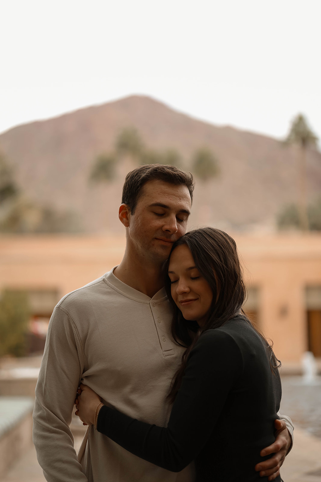 A couple smiling, with their eyes closed, while standing outdoors with palm trees and mountains in the background.
