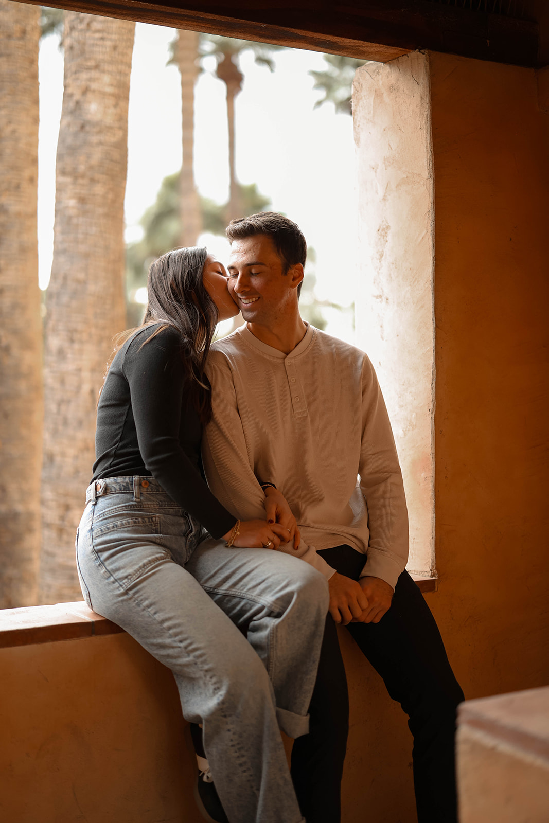 A sweet kiss on the cheek as the couple sits by a window, holding hands with palm trees in the background.