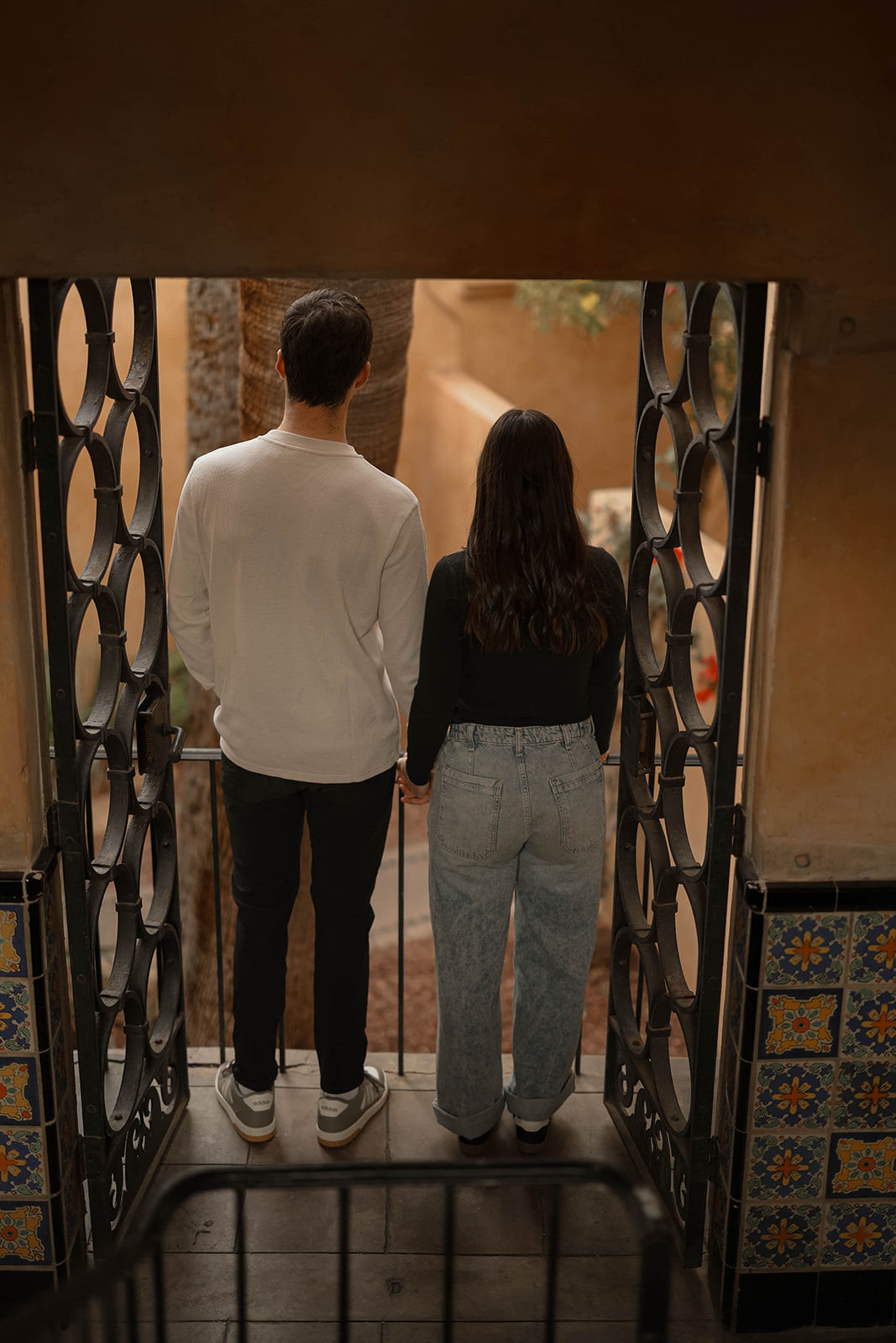 A couple stands hand in hand facing away from the camera, framed by ornate wrought-iron gates and colorful tile details.