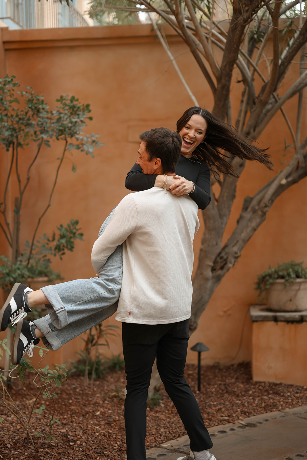A joyful moment of a man lifting his partner in the air as they laugh together near a courtyard tree.