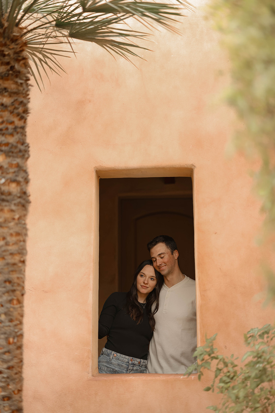 Two people rest together in a window cut-out of an adobe wall, framed by a palm tree.
