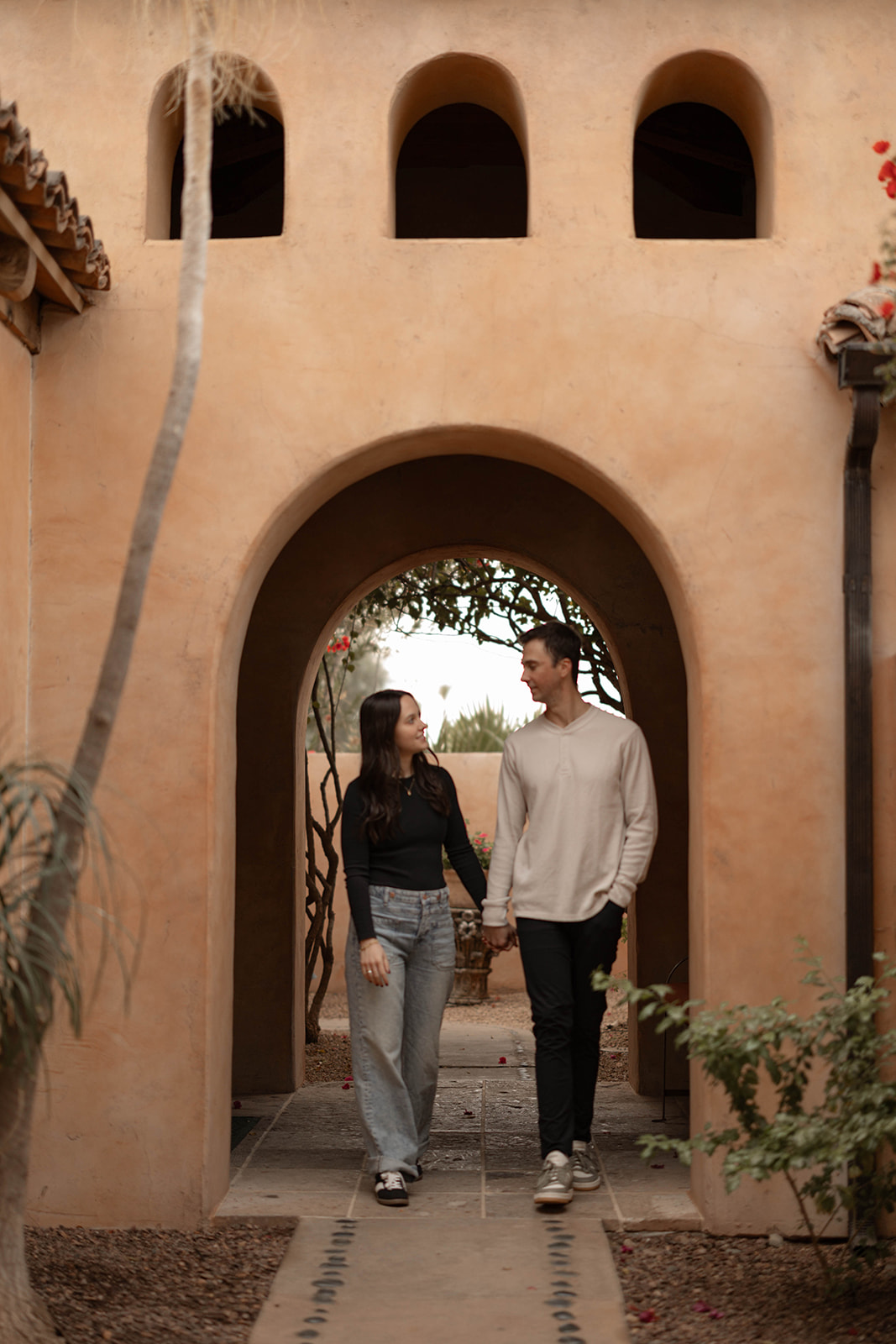 Warm couples photos walking hand-in-hand beneath an adobe archway.