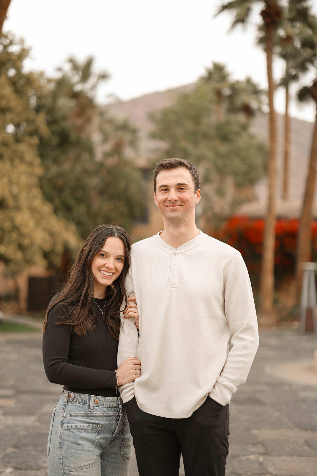 Smiling couple standing arm in arm outdoors with palm trees and a mountain view behind them.