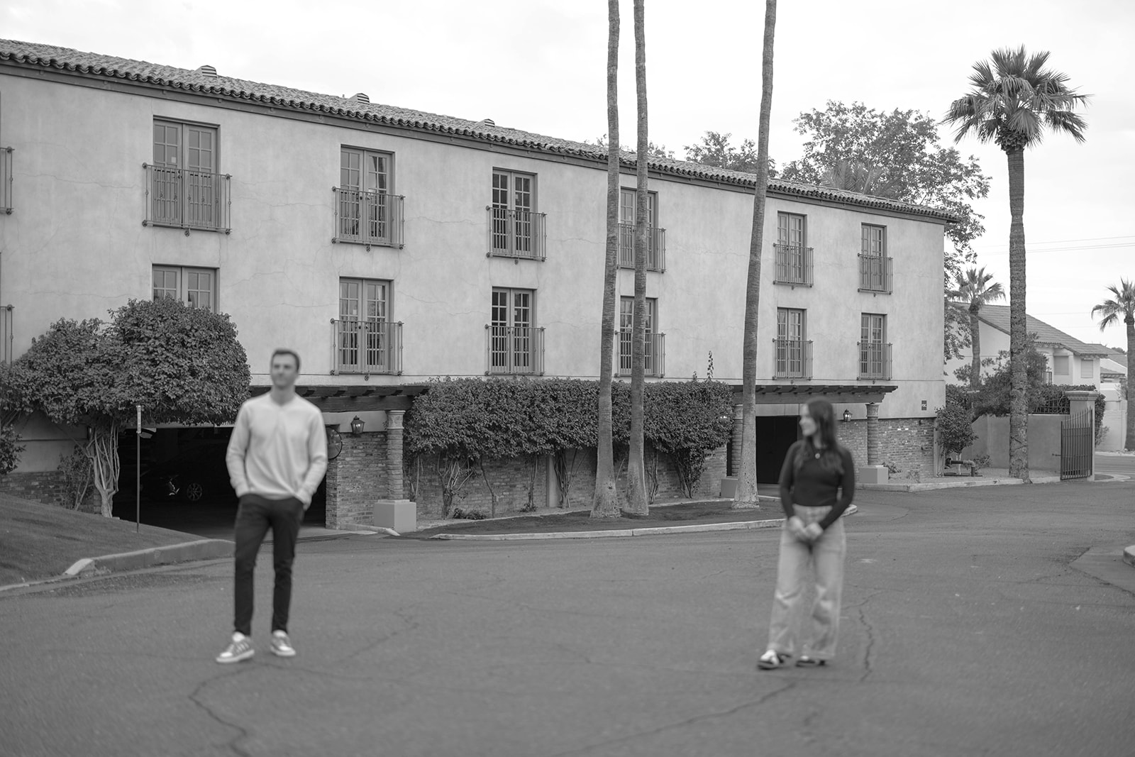 Black and white photo of a couple standing apart on a quiet street in front of a Spanish-style building.