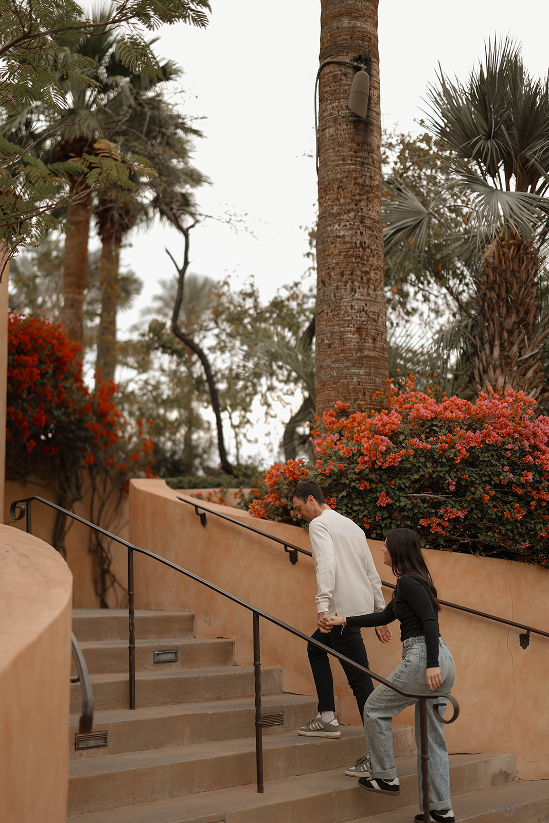 A man leads his partner up terracotta-colored stairs, surrounded by bright bougainvillea and palm trees.