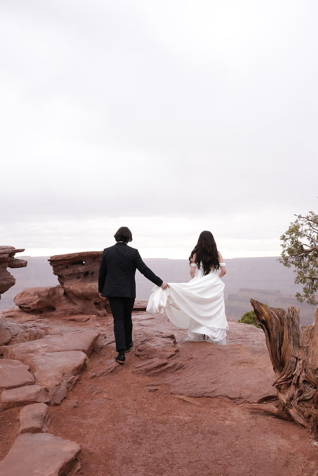 A couple walks hand-in-hand across a red rock cliff, the bride’s dress trailing behind her as the groom gently helps lift the fabric.