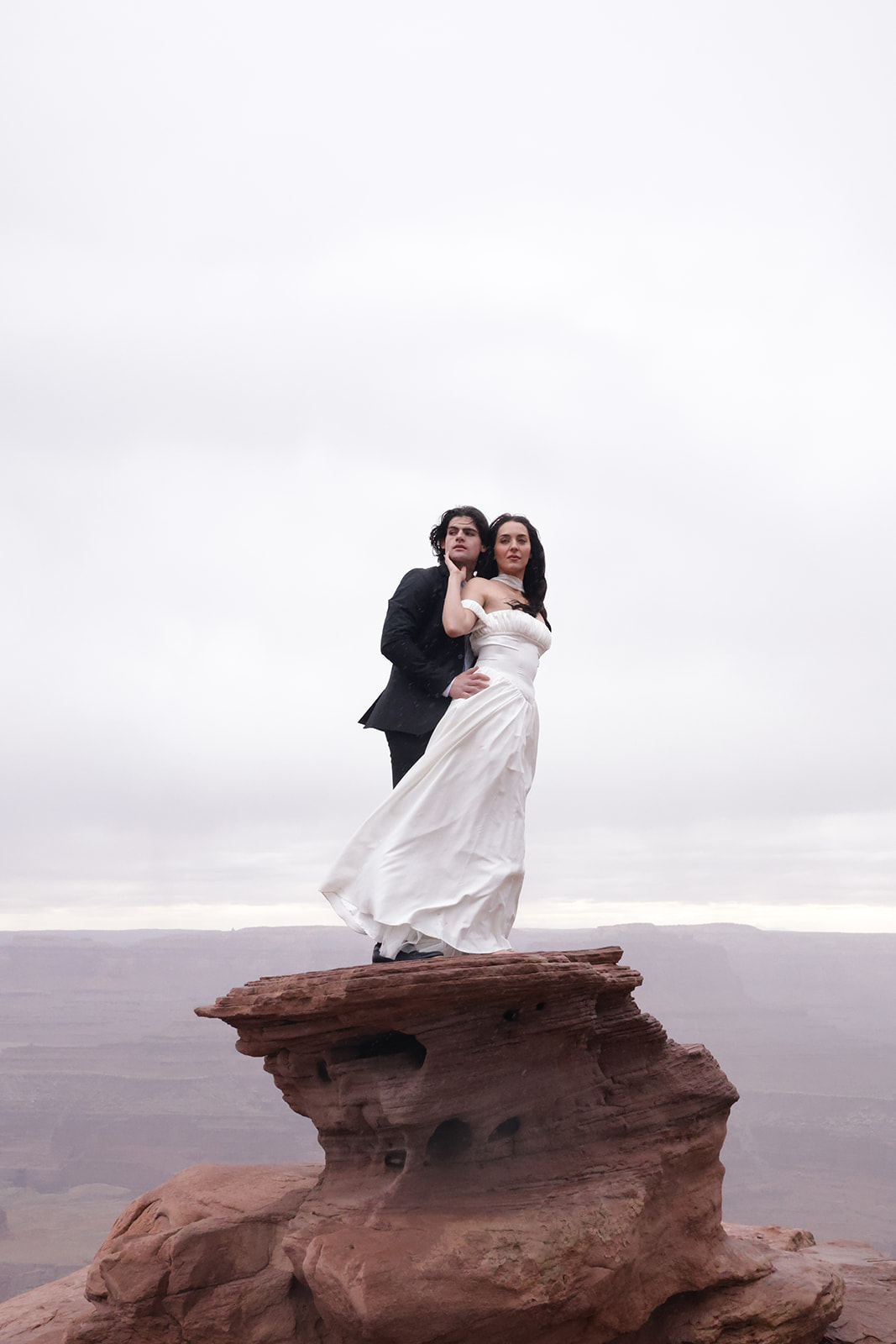 Bride and groom standing on red rock overlooking desert landscape, captured during a moody elopement session.