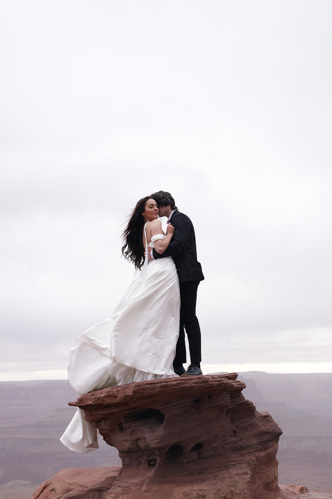 An intimate moment as a couple embraces on a dramatic rock ledge, wind catching the bride’s flowing elopement outfit against the backdrop of a misty canyon.