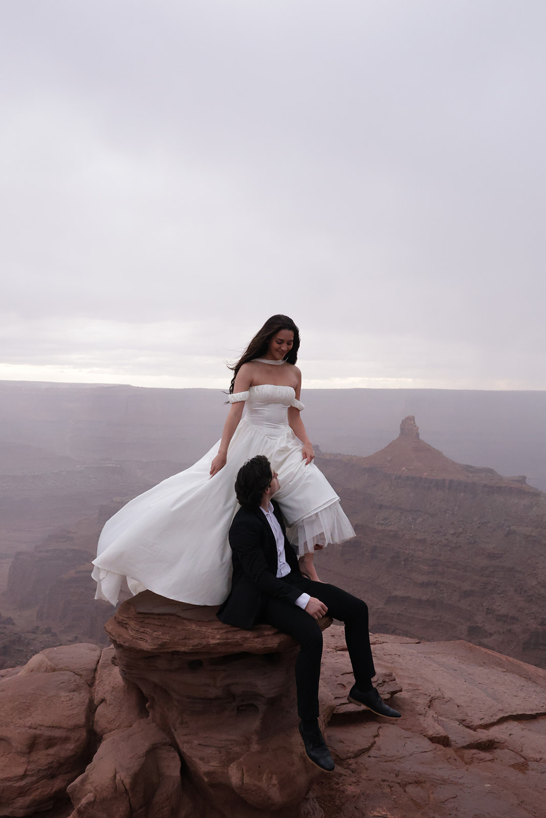 Bride and groom sharing a quiet moment on a red rock cliff, with the bride’s dress flowing in the breeze against the backdrop of the desert canyon.