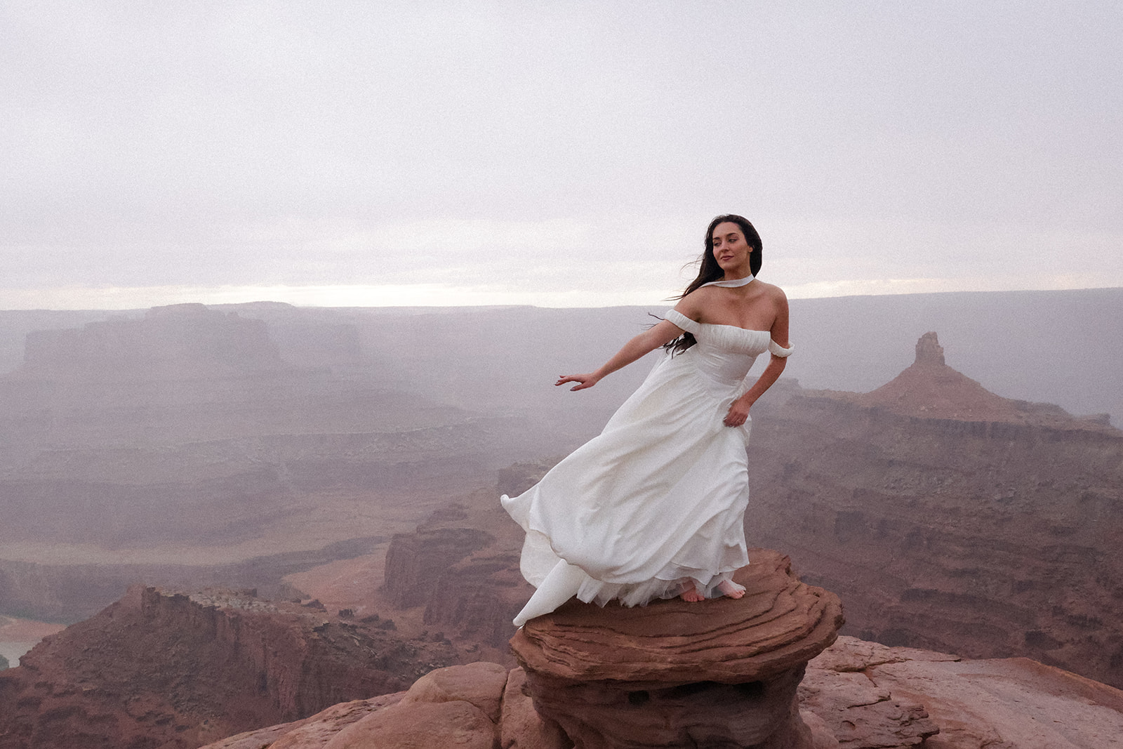 The bride stands barefoot on a red rock formation, her dress sweeping in the wind as she gazes over the canyon landscape.