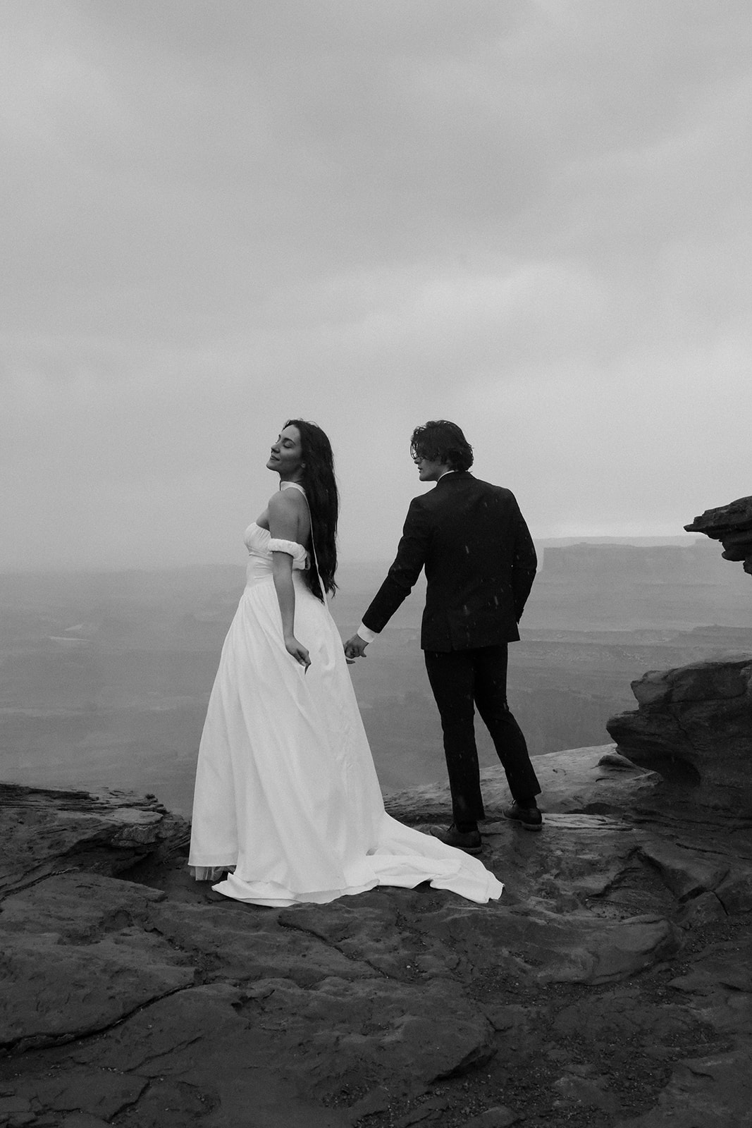 Bride in a flowy elopement outfit standing confidently on a cliff with expansive canyon views behind her.