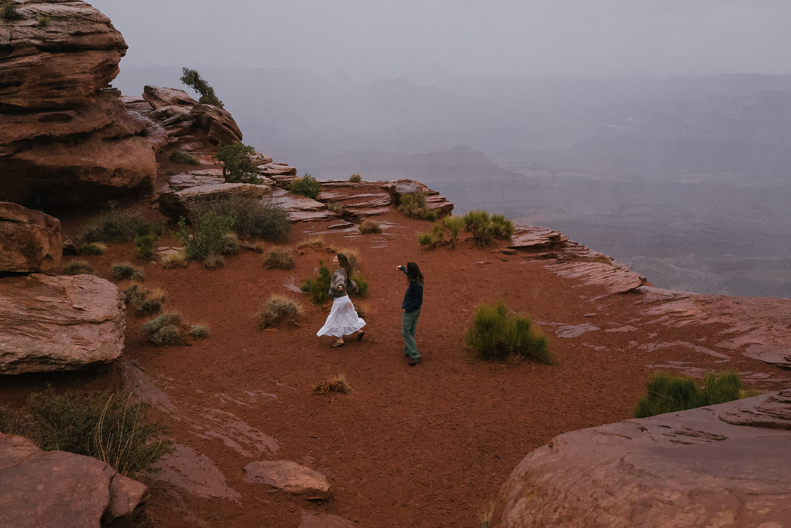 A behind-the-scenes moment capturing the bride walking through red desert terrain with her elopement outfit billowing slightly, guided by her groom.