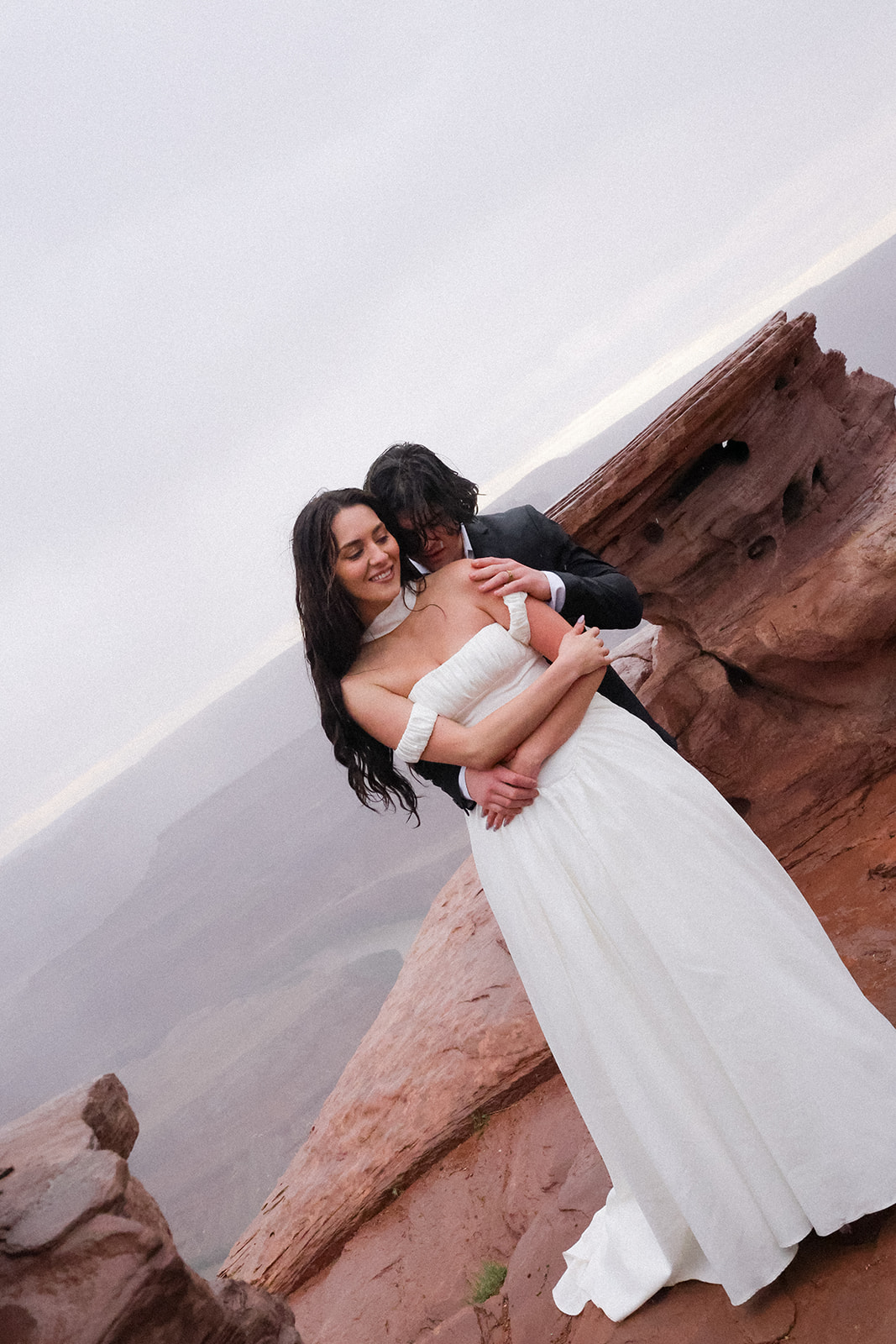 Couple wrapped in each other’s arms on a red rock ledge, with the bride’s elegant elopement outfit catching the light in the soft, misty air.
