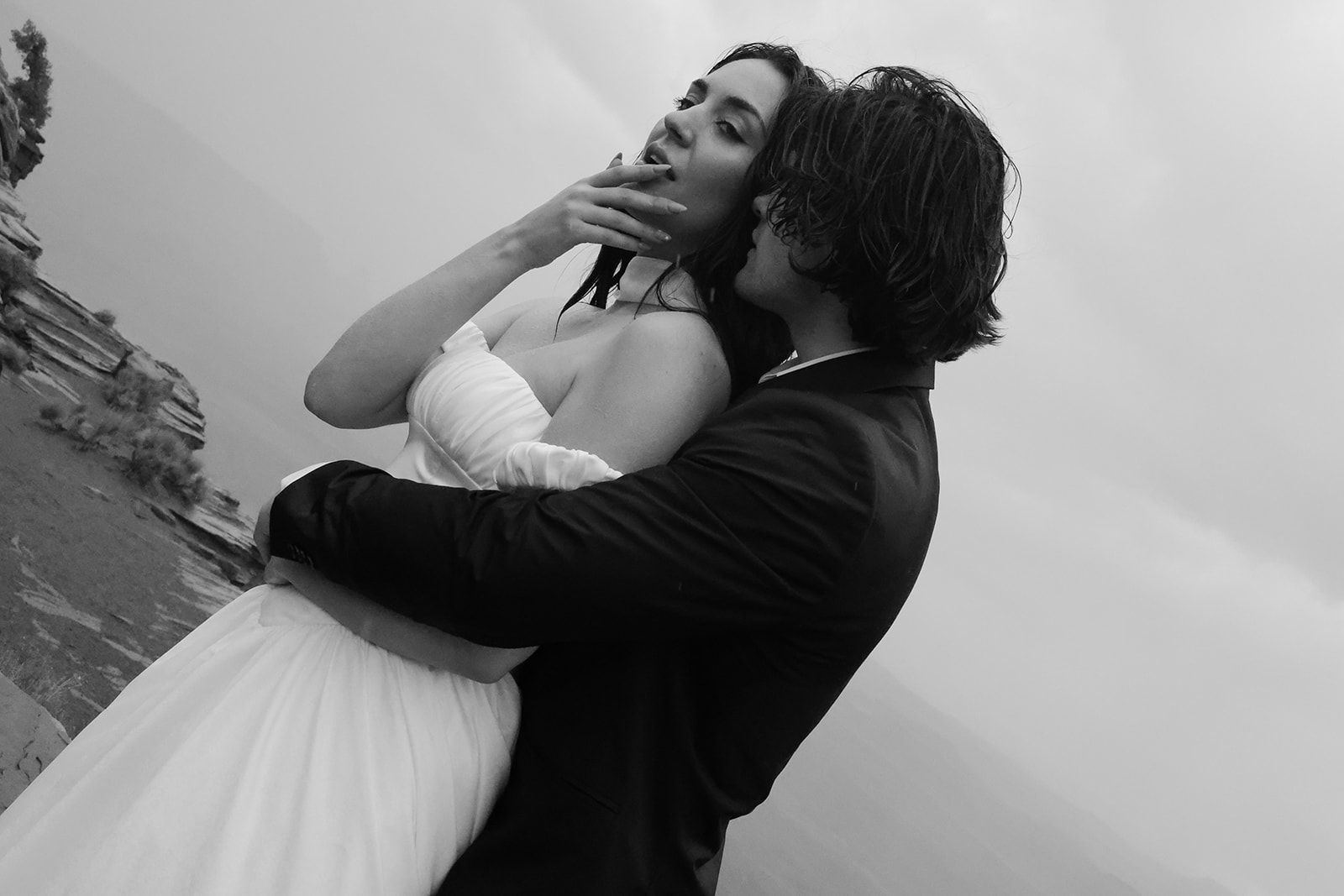 In a moody black-and-white portrait, the groom wraps his arms around the bride, her elopement outfit contrasting softly against the overcast sky.