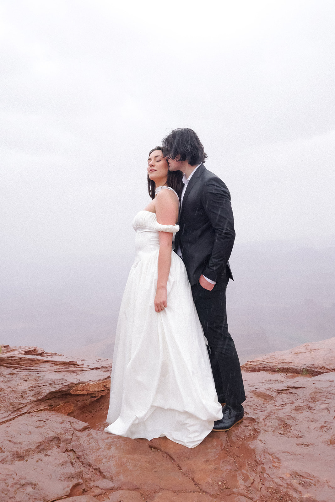 The couple shares a quiet, windswept moment at the canyon’s edge, the bride’s gown draping naturally over the rock beneath her.