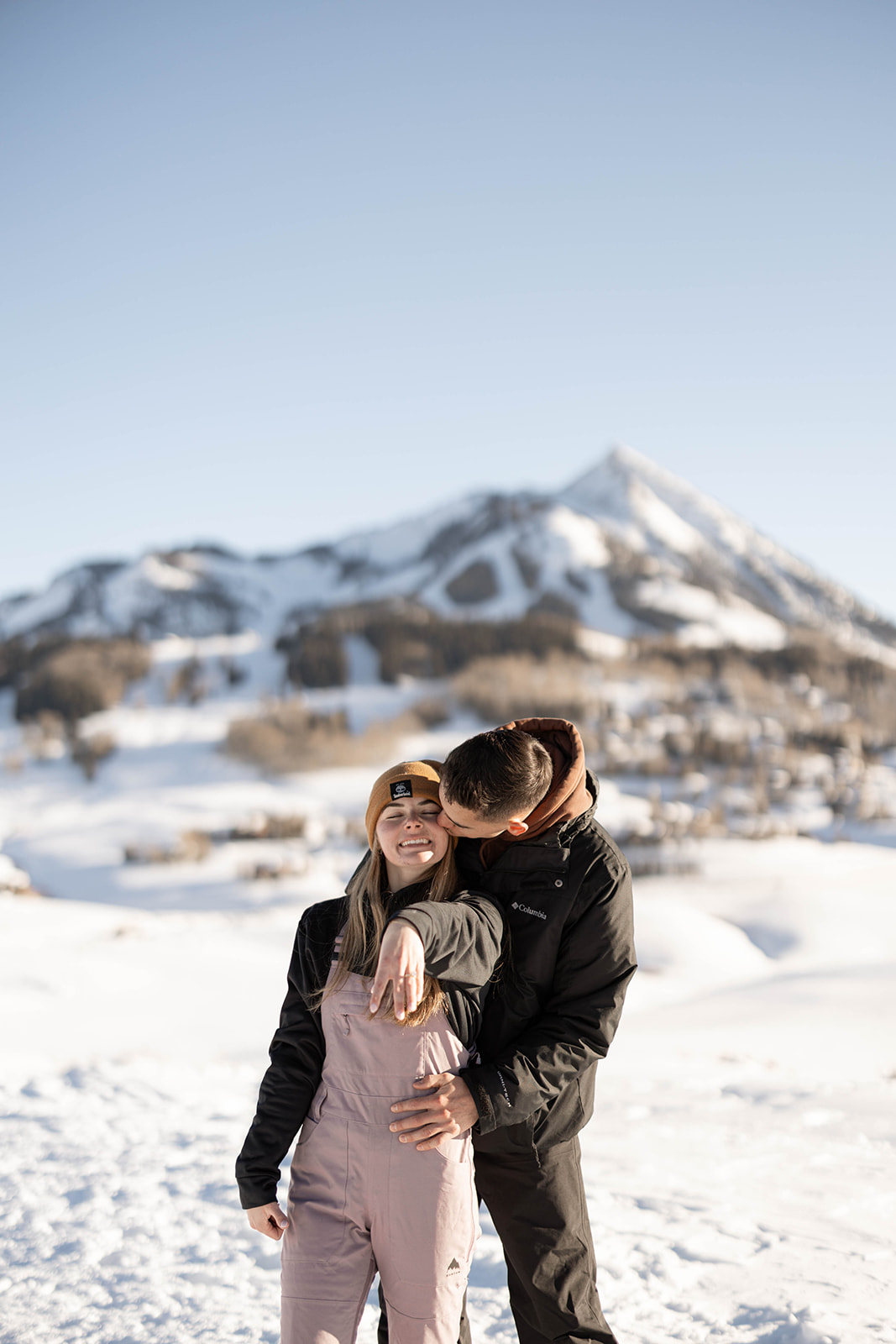 A newly engaged couple poses with the woman showing her ring, as her fiancé kisses her cheek against a mountain backdrop.