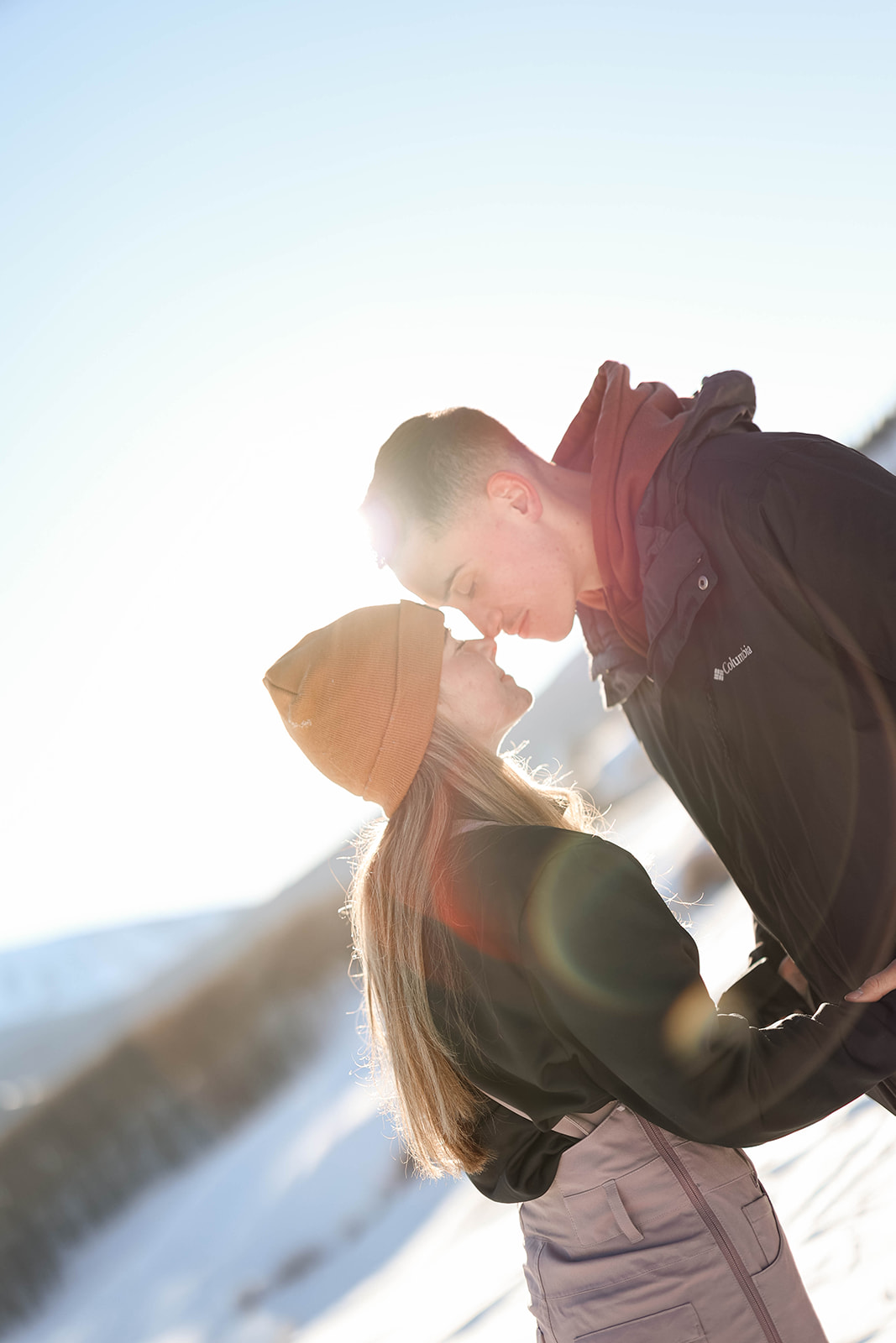 A couple shares a kiss on a snowy mountain slope, sunlight streaming behind them, creating a warm winter glow.
