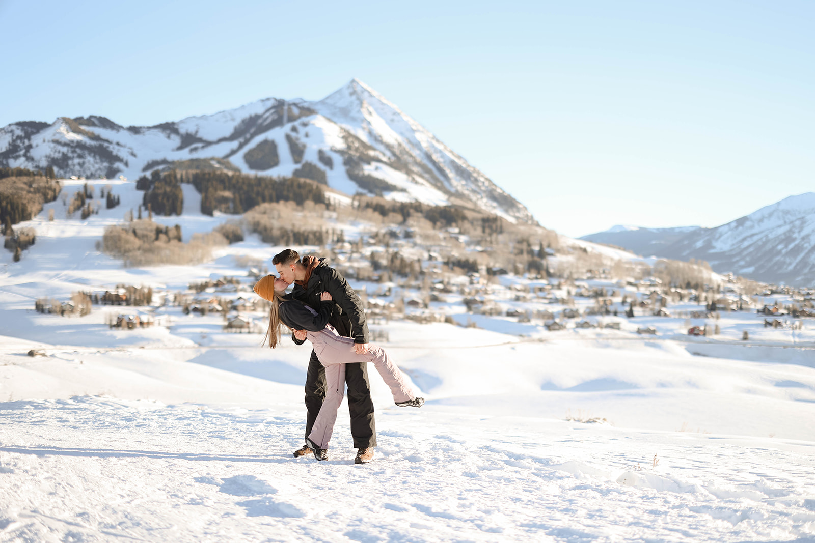 A man dips his partner for a kiss in the snow, the rugged mountain peaks towering behind them.
