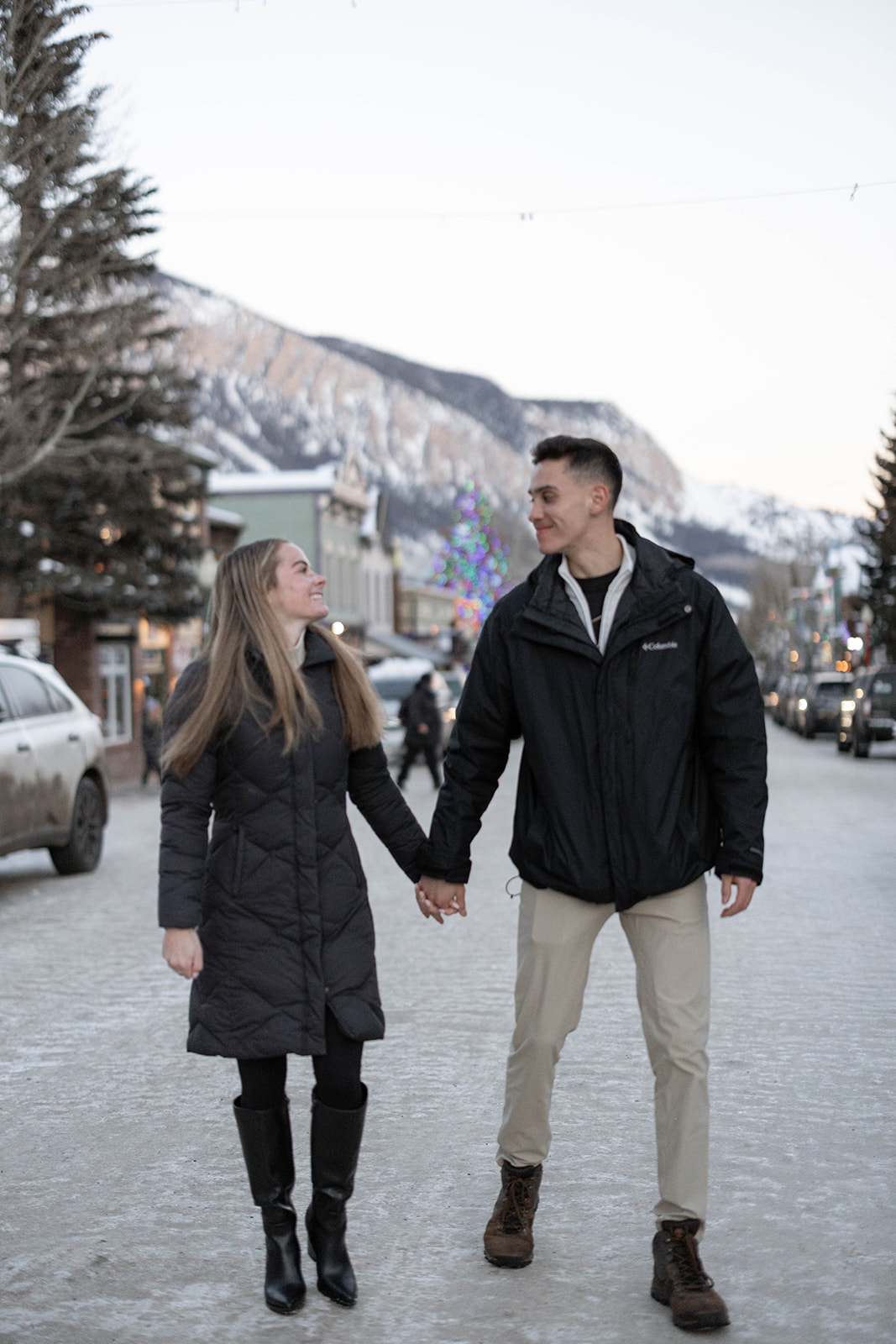 Couple holding hands and walking on a snow-lined street, smiling warmly at each other — surprise proposal happiness.
