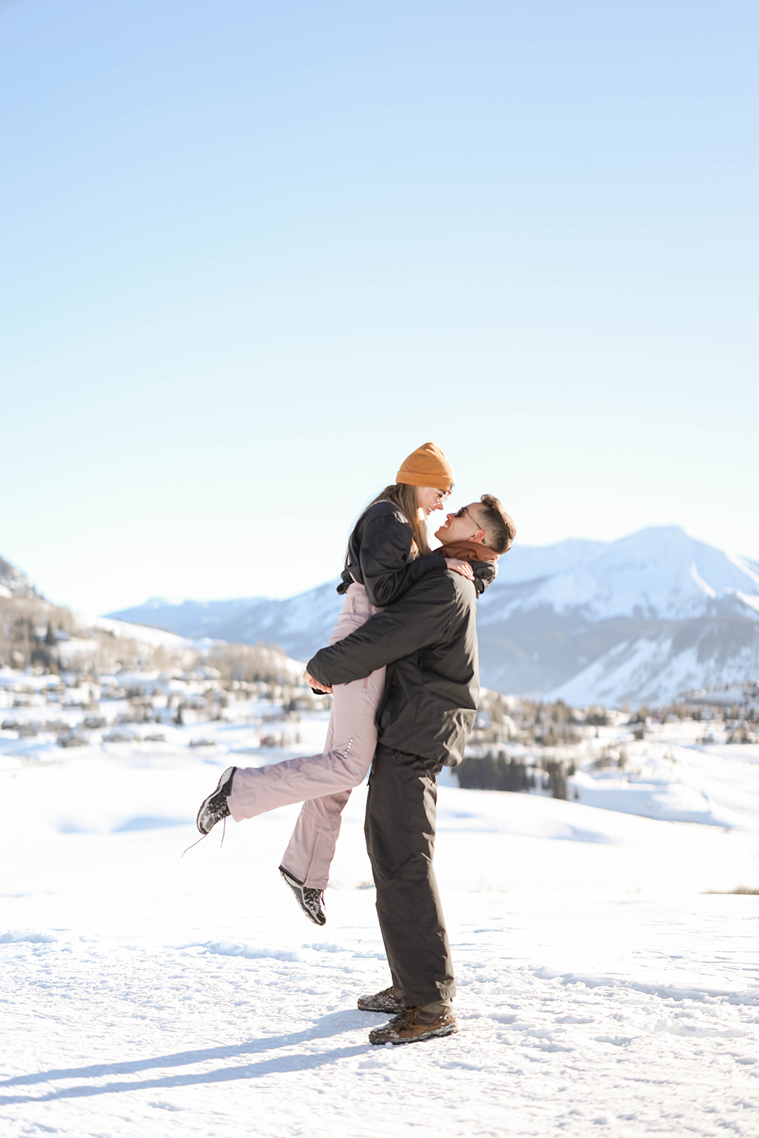 Man lifting his partner into the air during a joyful moment in the snow, mountains in the background, surprise proposal joy.