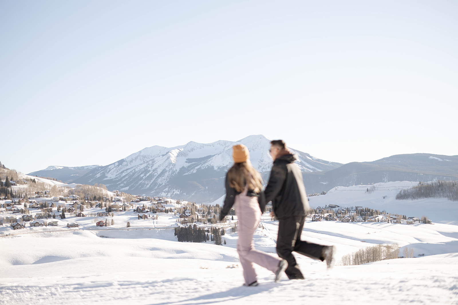 A couple walks hand in hand across a snowy hill, bundled in winter gear, with Crested Butte’s snow-covered peaks and a mountain village in the background during a surprise proposal.