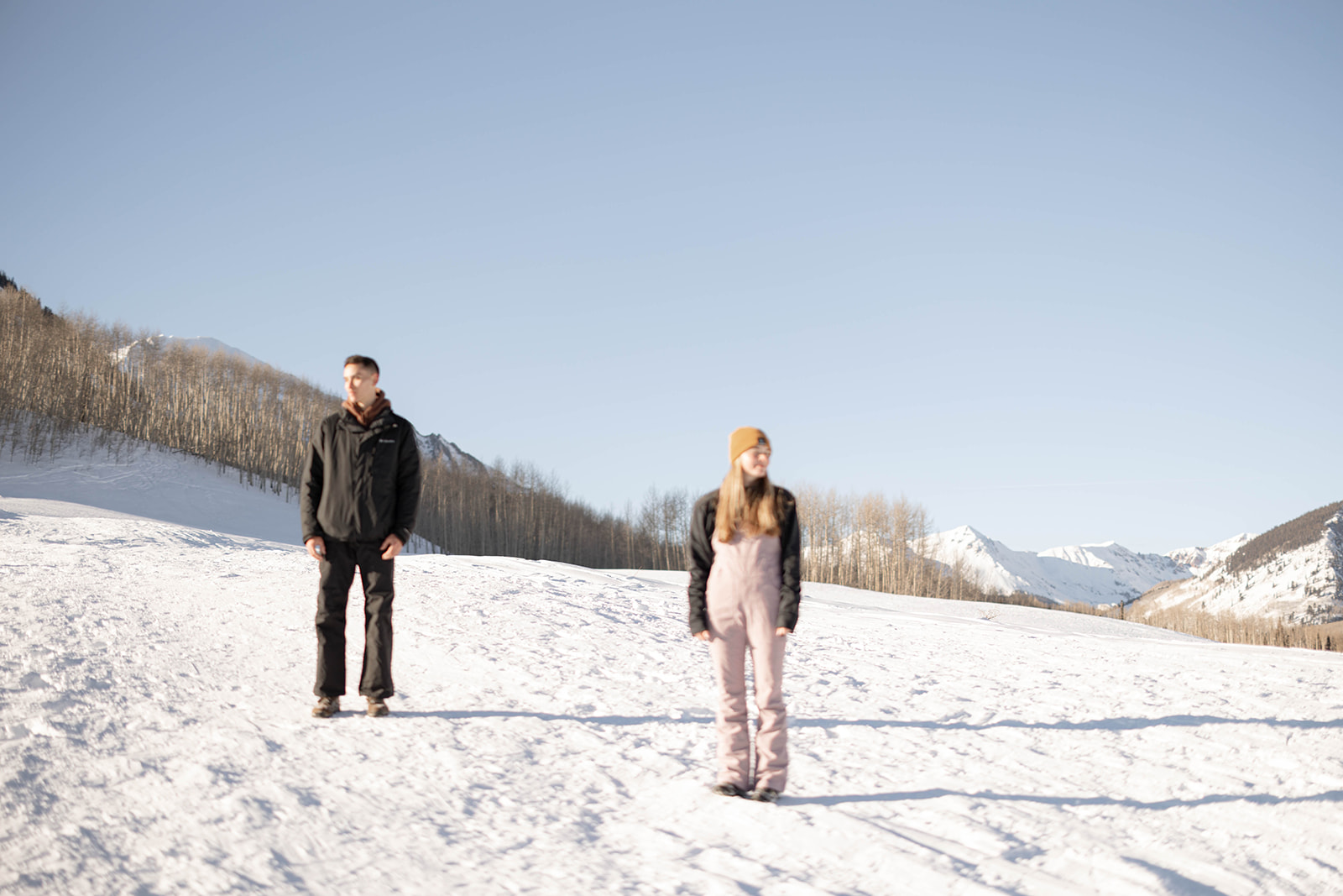 Couple standing apart on a snowy mountain slope with bare trees and a clear blue sky in the background.