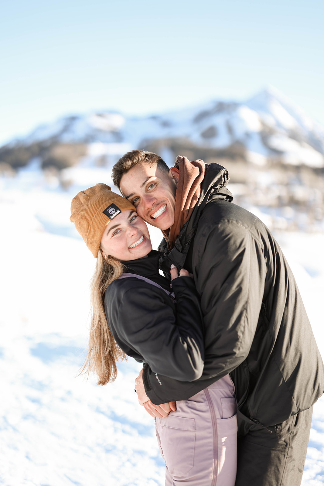 Couple embracing in the snow with mountains behind them, both smiling at the camera.
