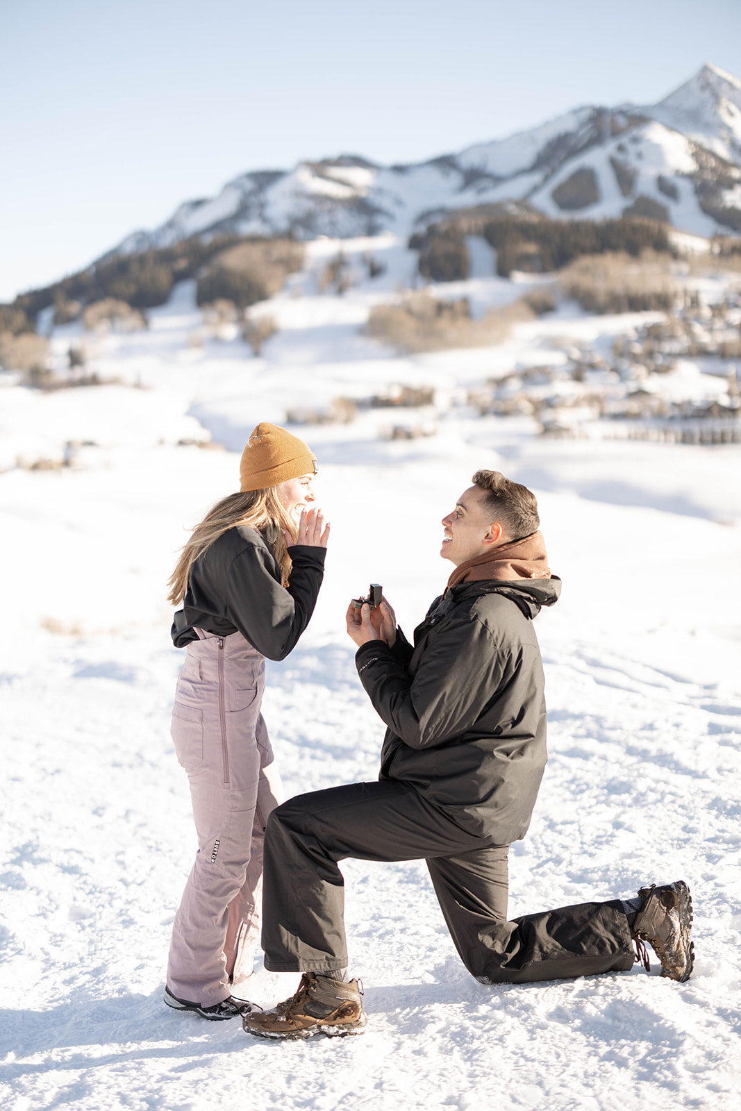 A man kneels in the snow, holding out a ring box, as his partner reacts joyfully to the surprise proposal.