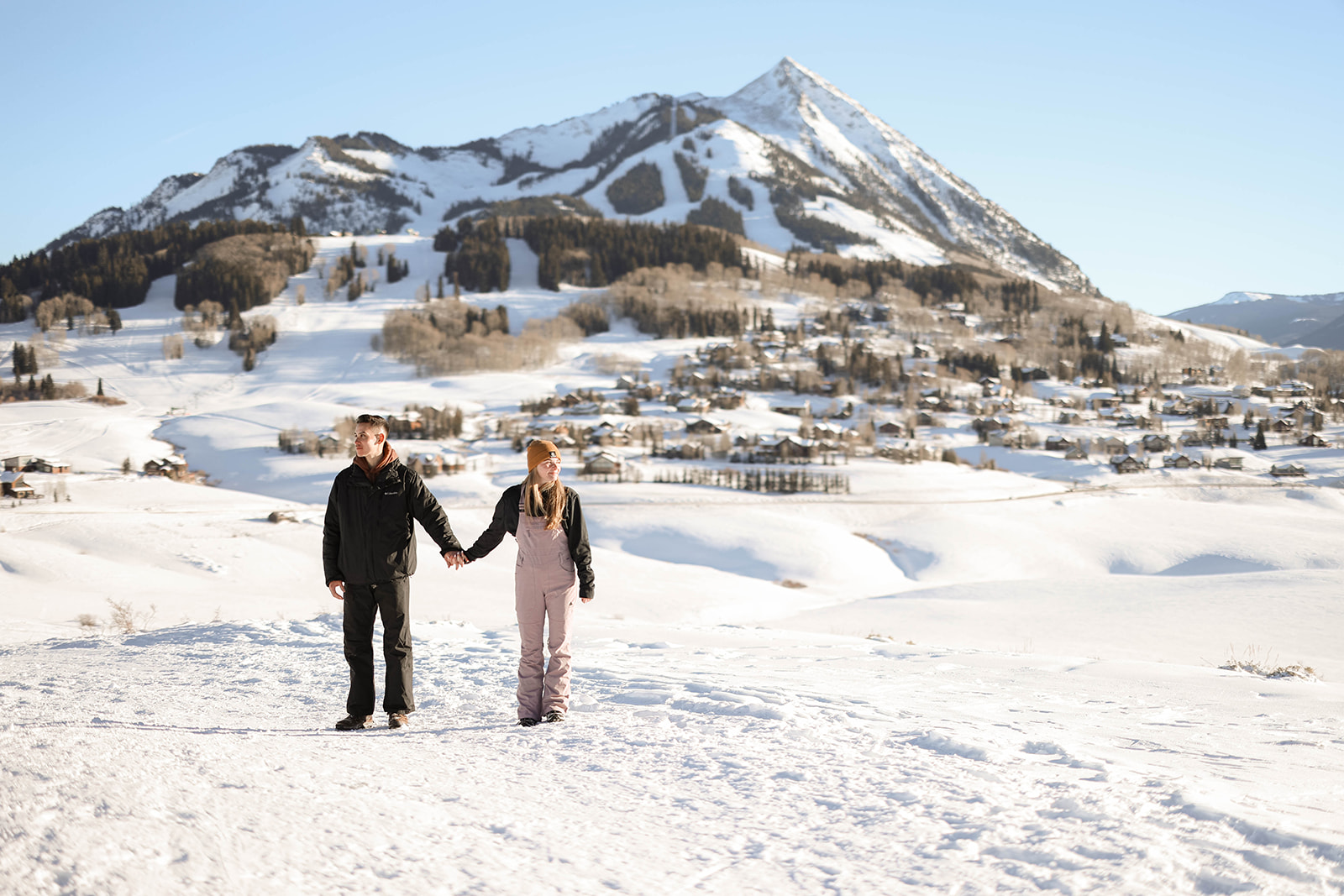 A couple holds hands, standing against a panoramic snowy mountain view under clear blue skies.
