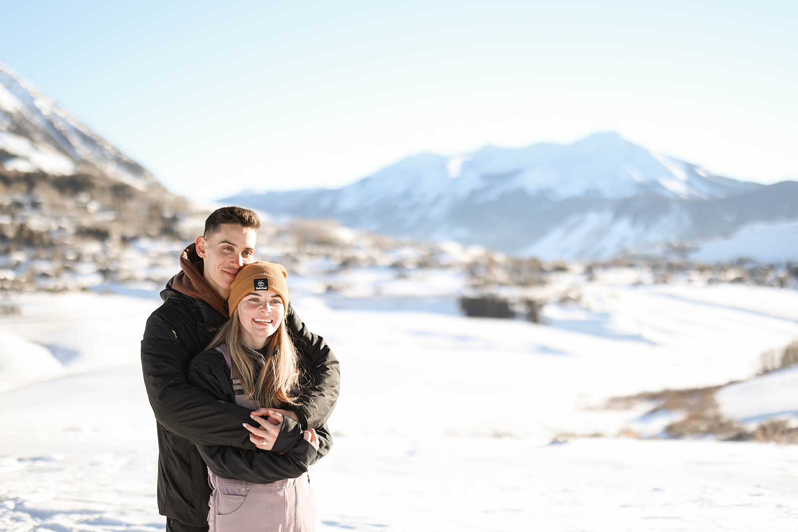 A man embraces his partner from behind, both smiling warmly with a snow-covered valley in the background during their surprise proposal.