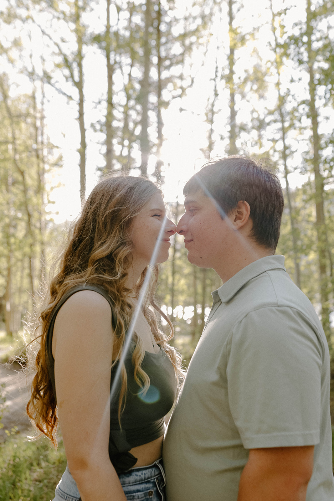 Sweet moment of a couple standing nose-to-nose in the forest with soft sunlight filtering through the trees.