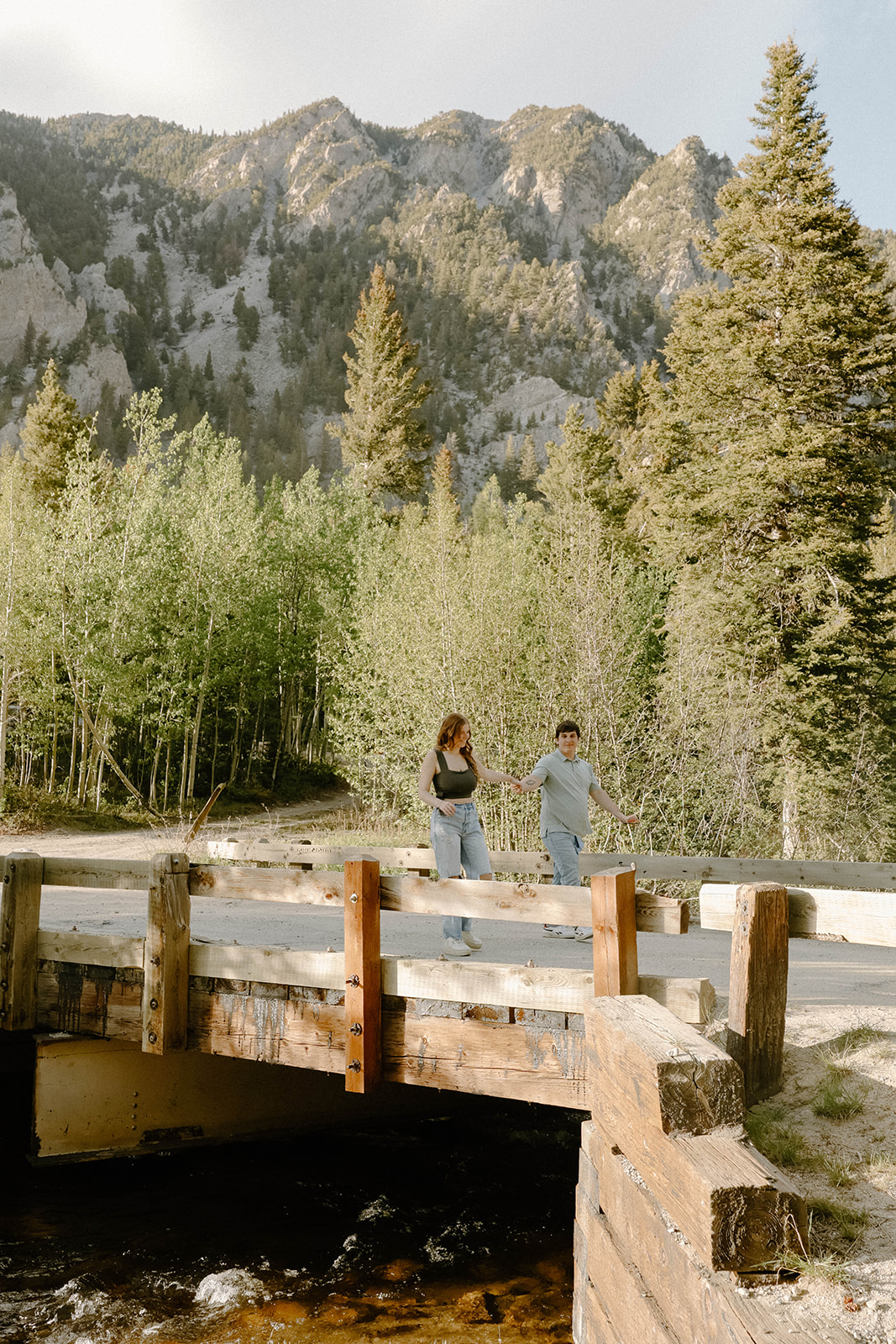 Couple walking together across a wooden bridge with pine trees and rugged mountain cliffs in the distance.