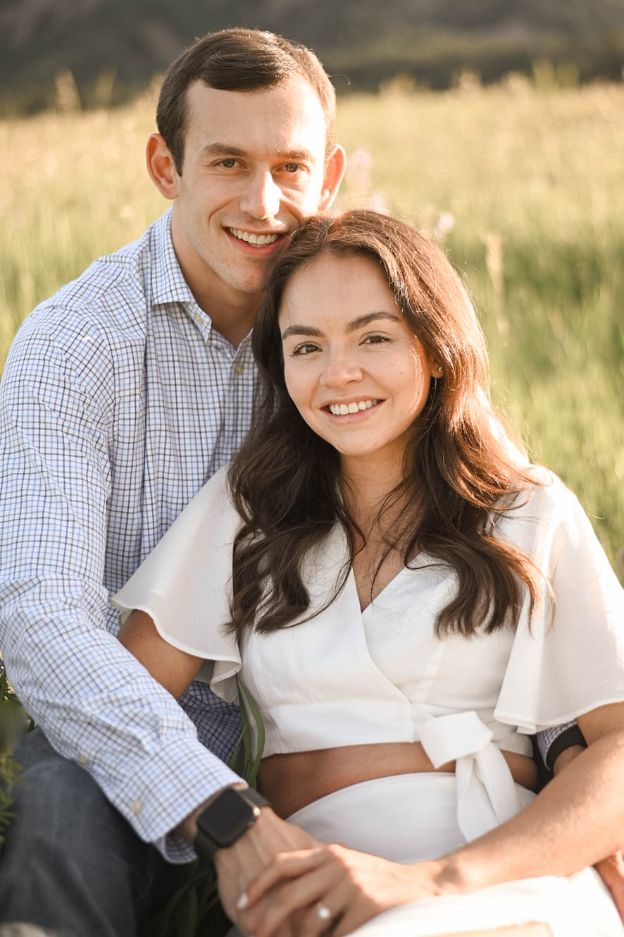 Engaged couple sitting close together in a sunlit meadow at Chautauqua Park, Boulder, smiling at the camera.