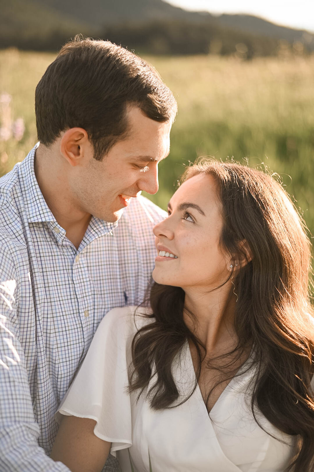 Engaged couple sitting close in tall grass, smiling and gazing at each other in golden sunset light.