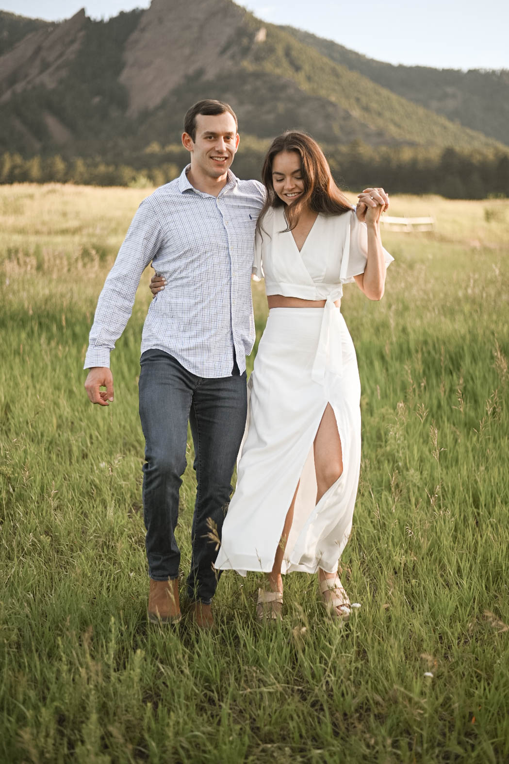 Couple walking arm in arm through a grassy field at Chautauqua Park, Boulder, with the Flatirons rising in the background.