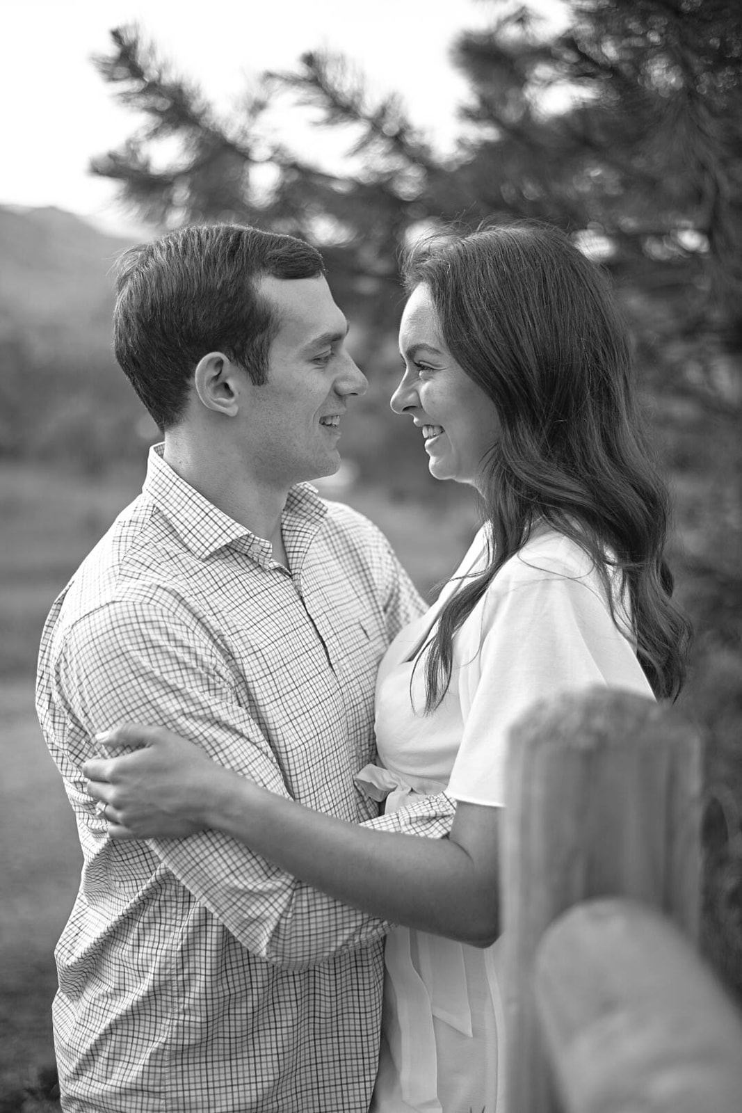 Black and white photo of a couple smiling at each other, leaning close with trees softly blurred in the background.