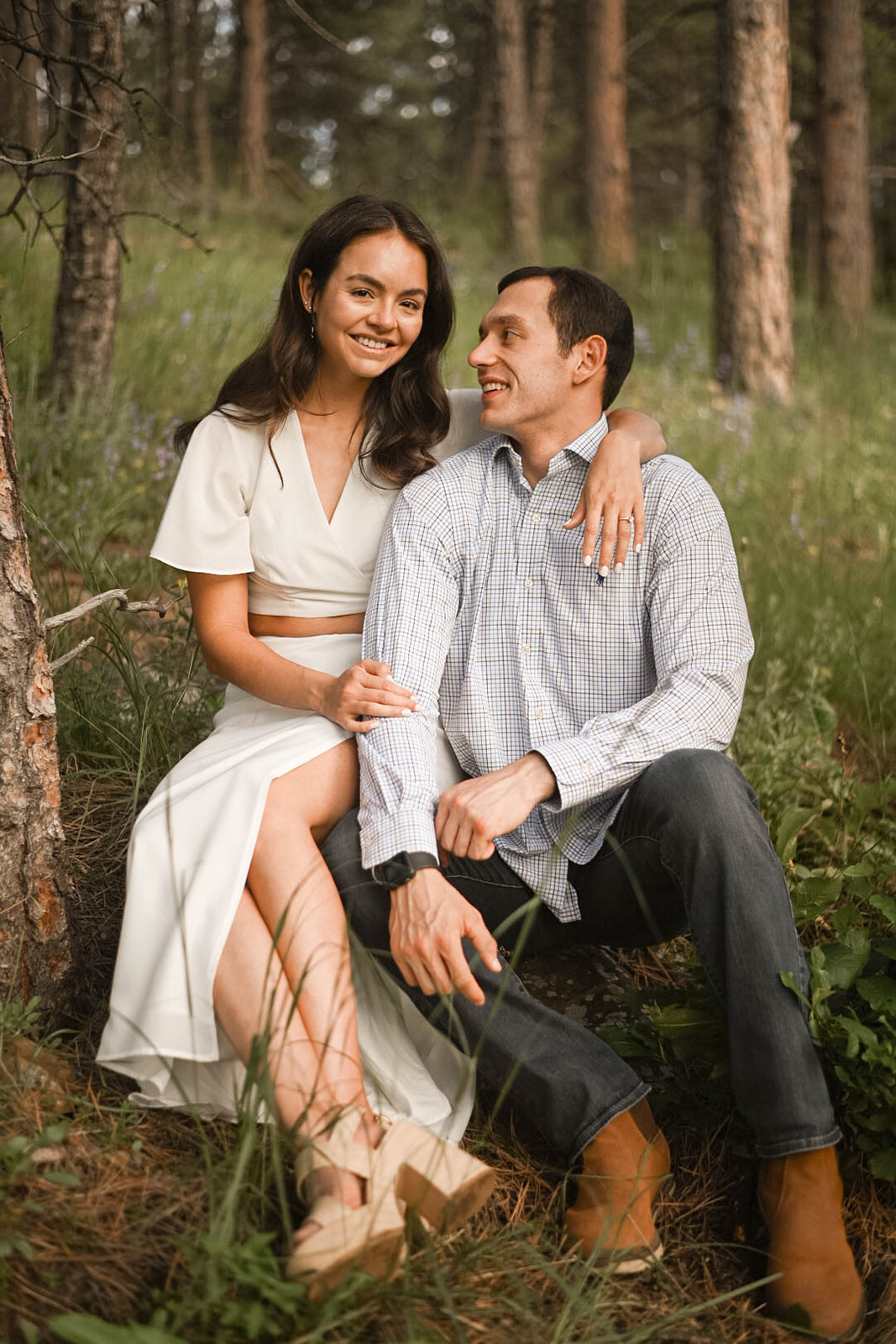 Woman in a white dress sitting beside her partner in a forest clearing, both smiling and leaning close.